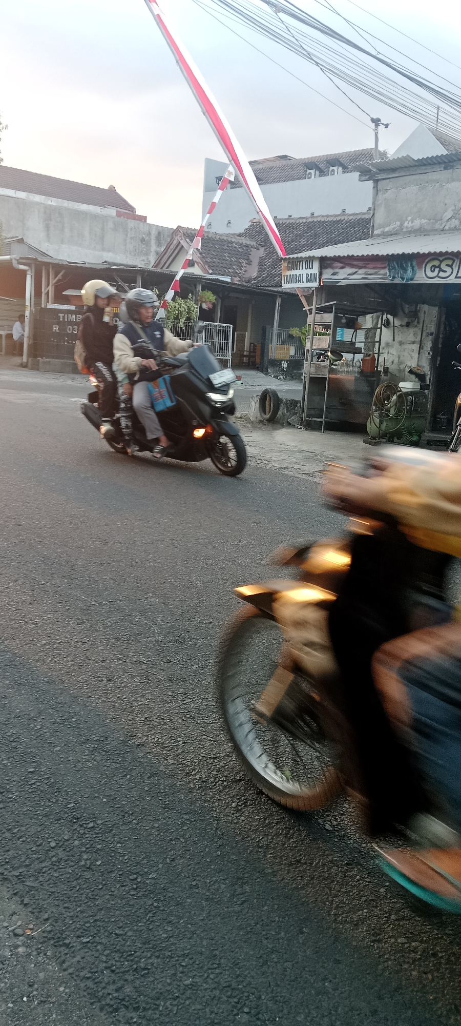 In Maguwoharjo, Indonesia, a scene unfolds on a paved road, possibly near a residential or commercial area, indicated by buildings in the background. The sky is overcast, with hints of dawn or dusk light. A red and white Indonesian flag, likely a banner, is prominently displayed, stretched across poles and wires overhead.
The foreground is dominated by a black scooter with a tinted windshield, its front wheel and fender blurred due to motion. A person, whose body is largely obscured by motion blur and a yellow jacket, is riding this scooter. They appear to be wearing a helmet.
Further back on the road, a second scooter is visible, also in motion. This scooter is black and carries two individuals. The driver, wearing a helmet, is looking forward. The passenger, behind the driver, is also wearing a helmet and is looking to their left. The passenger is carrying a blue bag.
The environment includes a gray asphalt road with some white lines and a tire lying on the side. Buildings lining the road appear to be shops or houses with tiled roofs. One building has a sign that reads "GANTI OLI TAMBAL BAN", which translates to "Oil Change Tire Patch". Another sign partially visible on a building reads "GS". The overall impression is a typical street scene in a populated Indonesian area.