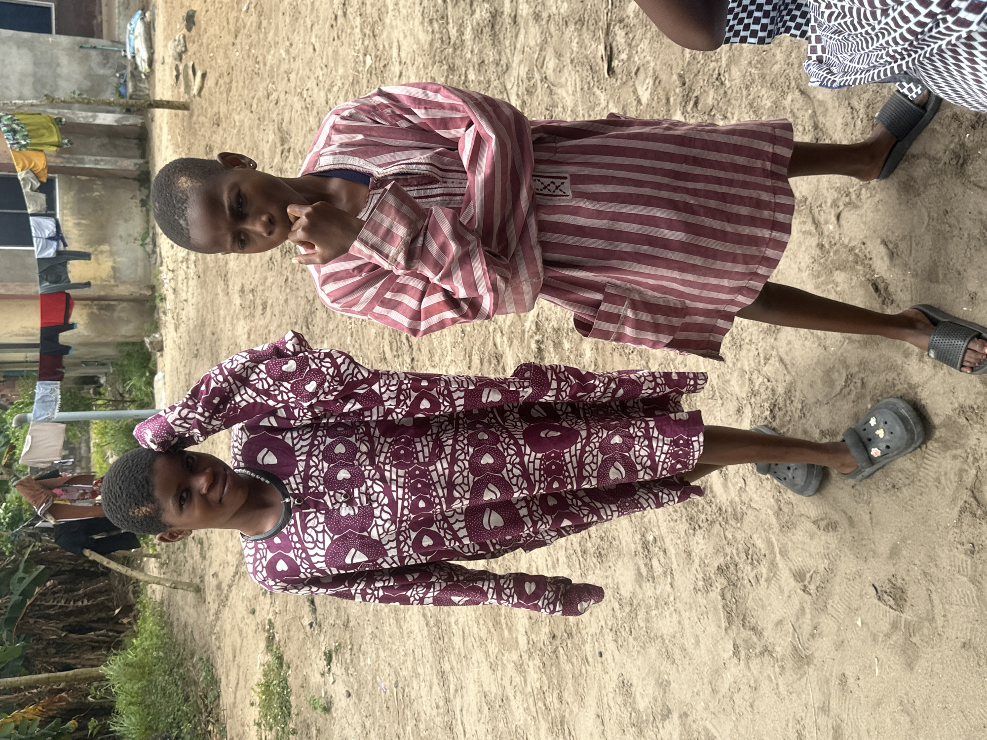 Two young individuals are standing on a sandy ground in what appears to be a residential area in Badagry, Nigeria. The individual on the left, facing slightly towards the viewer, wears a long, dark purple robe with a white patterned design and grey Crocs. This person has their left arm raised, with their hand resting on their head. The individual on the right, facing away from the viewer, wears a striped pink and white robe and black slides. Their right hand is near their mouth, and they are looking down. In the background, laundry is hanging on a clothesline strung between two poles. Various structures, including a building with windows and a rough fence, are visible. The ground is a mixture of sand and some sparse vegetation.