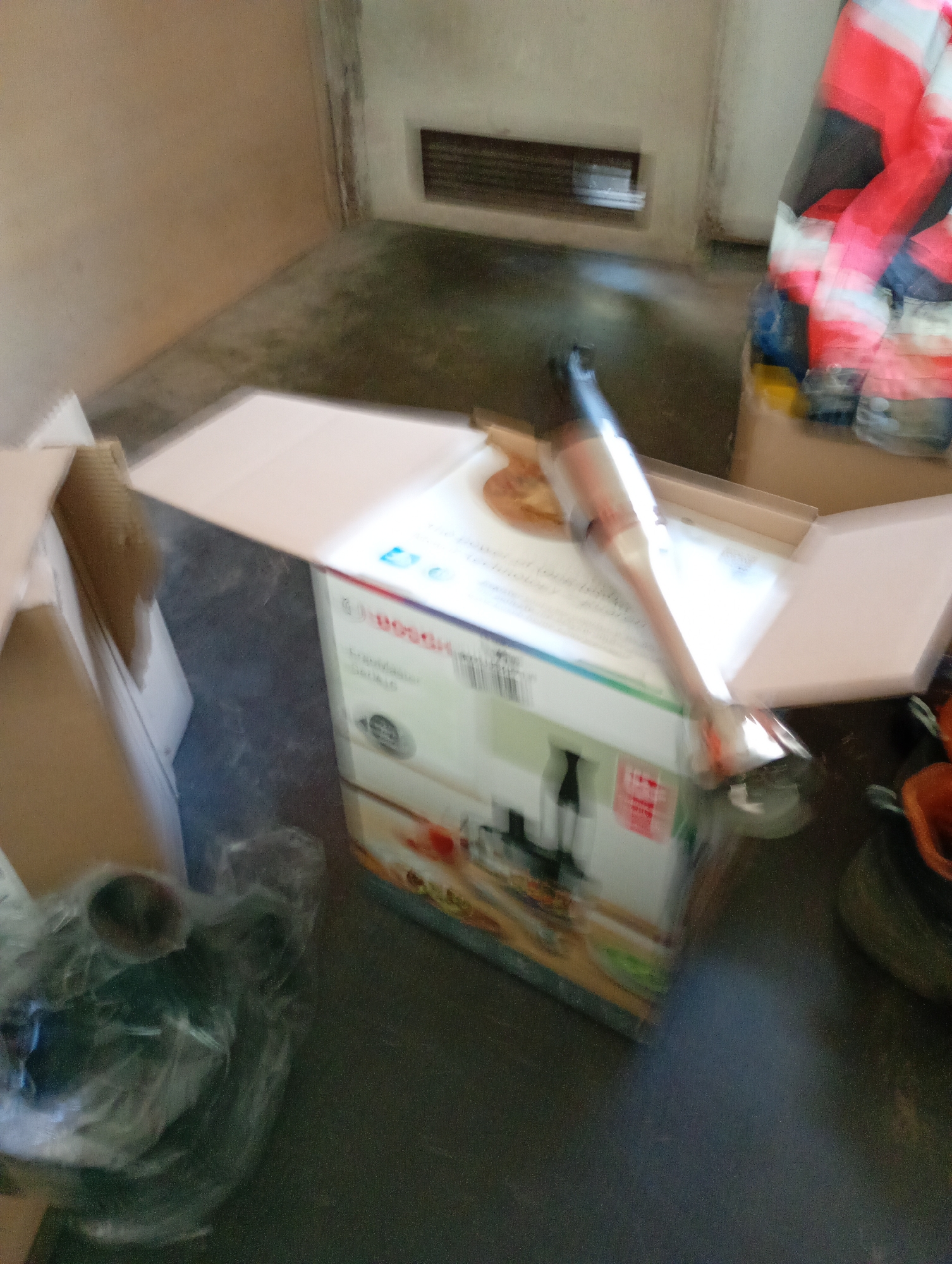 The image depicts an indoor scene, possibly a storage area or a room with concrete flooring. A white and colorful cardboard box, likely containing an appliance, is prominently placed. An immersion blender is visible, partially inside the box and extending outwards. To the right of the box, there are items that appear to be clothing, possibly workwear with red and white reflective stripes. Further to the right, a brown plastic container is partially visible. In the foreground on the left, a dark object wrapped in plastic, possibly another item or debris, is present. The overall environment is dimly lit and appears somewhat cluttered. No specific landmarks or contextual clues are present to determine a location within Białystok, Poland.