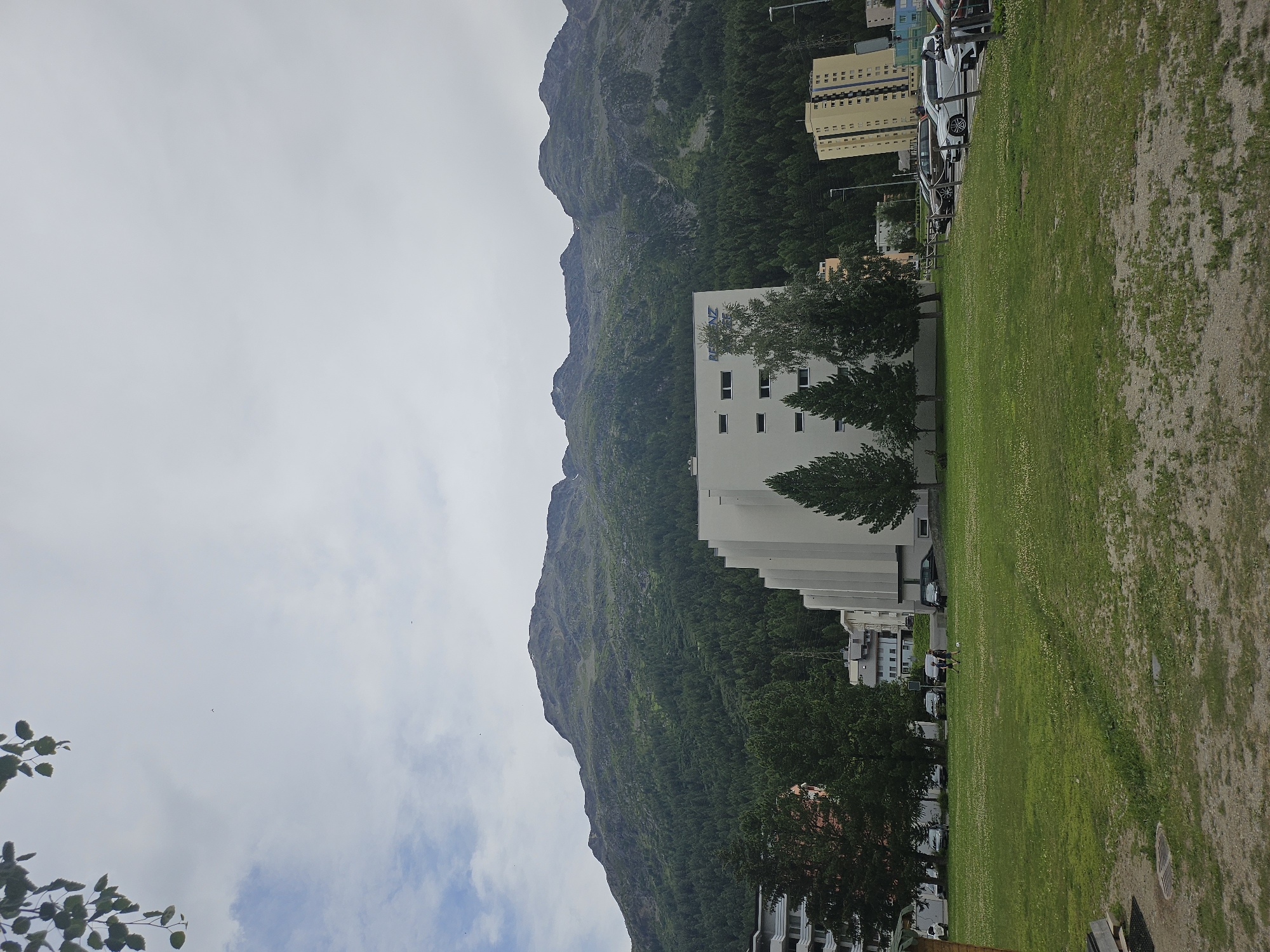 In St. Moritz, Switzerland, a modern white building with a prominent blue logo on its facade stands in the foreground, partially obscured by trees. This building is situated across from a lush green lawn where two individuals are playing soccer. Beyond the lawn, several cars are parked in a lot adjacent to another modern yellow building. The backdrop of the scene is dominated by a vast, verdant mountain range, its slopes densely covered with evergreen trees. The sky above is overcast with scattered clouds.