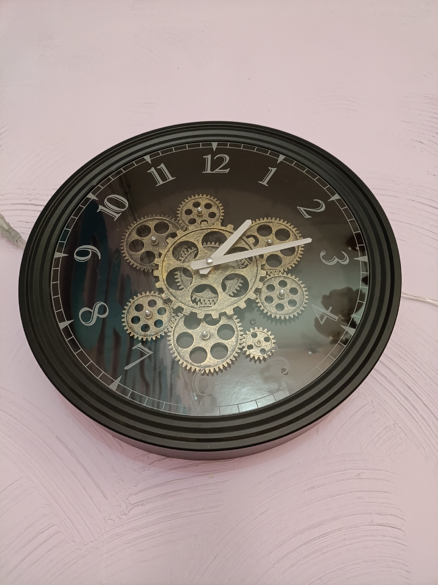 The image shows a round, black wall clock with a transparent face displaying a set of gold-colored gears in the center. The clock face has silver numbers and markers. The time displayed on the clock is approximately 11:16. The clock is mounted on a pale pink wall with visible textured paint. There are no people visible. The location is unknown.