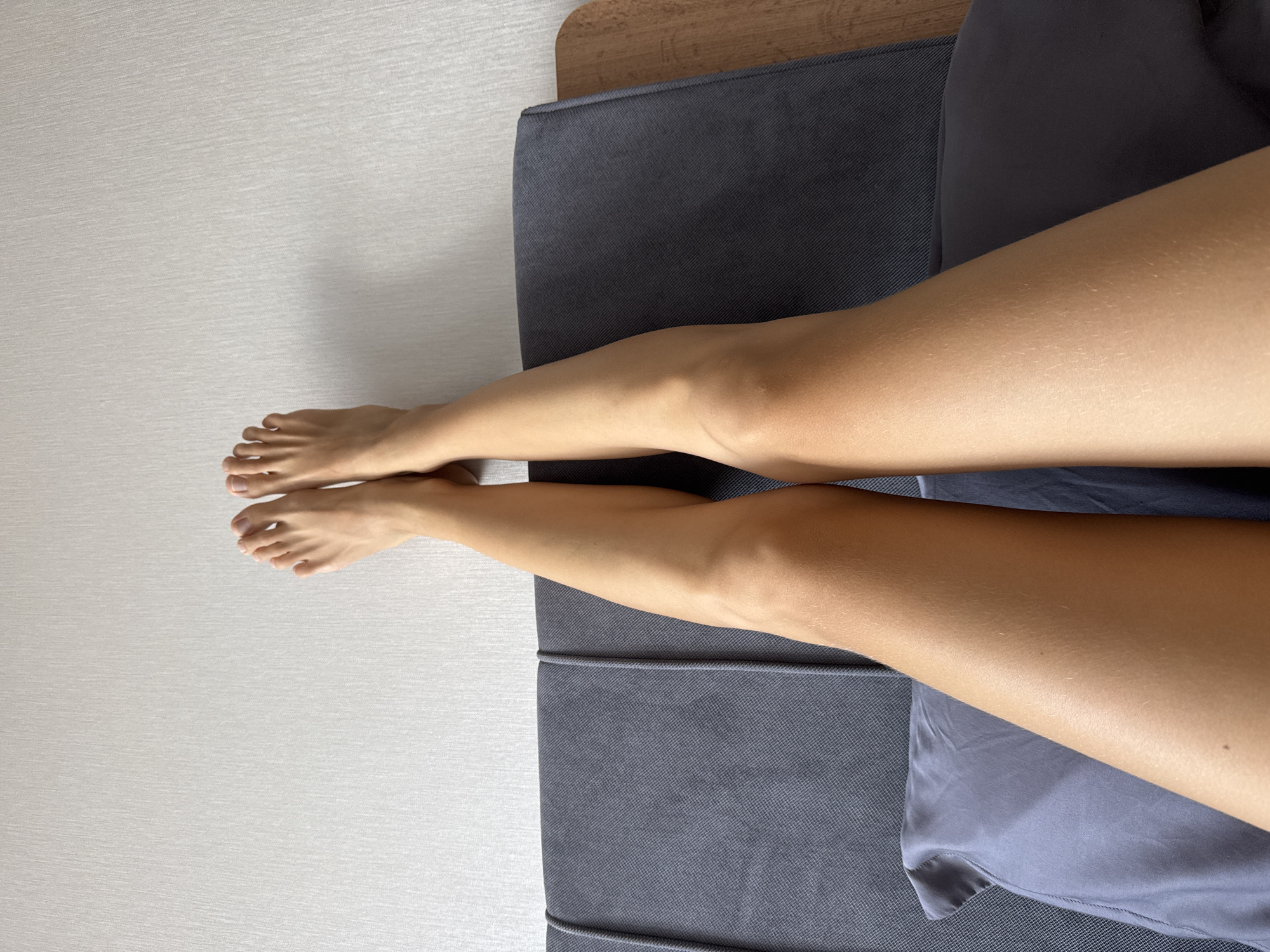 The image shows a top-down view of a person's legs resting on a dark gray sofa. The legs are tanned and positioned diagonally across the frame, with the feet pointing towards the upper left corner. The sofa is placed against a light gray wall. A portion of a wooden headboard is visible at the top center of the frame. The location is Unknown, Unknown.