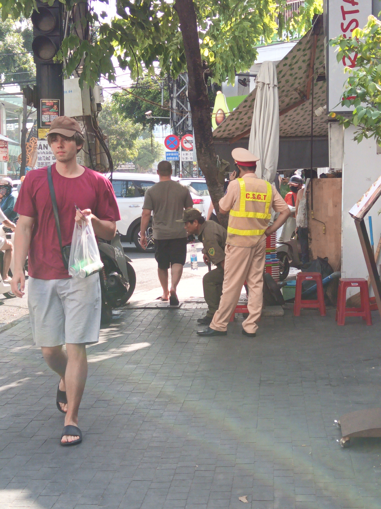 A man in a burgundy t-shirt and light grey shorts walks on a paved sidewalk carrying a clear plastic bag with two green cans inside. He is wearing black sandals. To his left, a traffic signal with three lights is visible, along with trees and other foliage. Behind him, a man in a tan uniform with a reflective yellow vest is standing, and another man in a tan uniform is sitting on a red stool. Further in the background, a white car and a motorcycle are parked. The scene appears to be in an urban environment, possibly near a street or intersection, with commercial establishments visible in the distance.