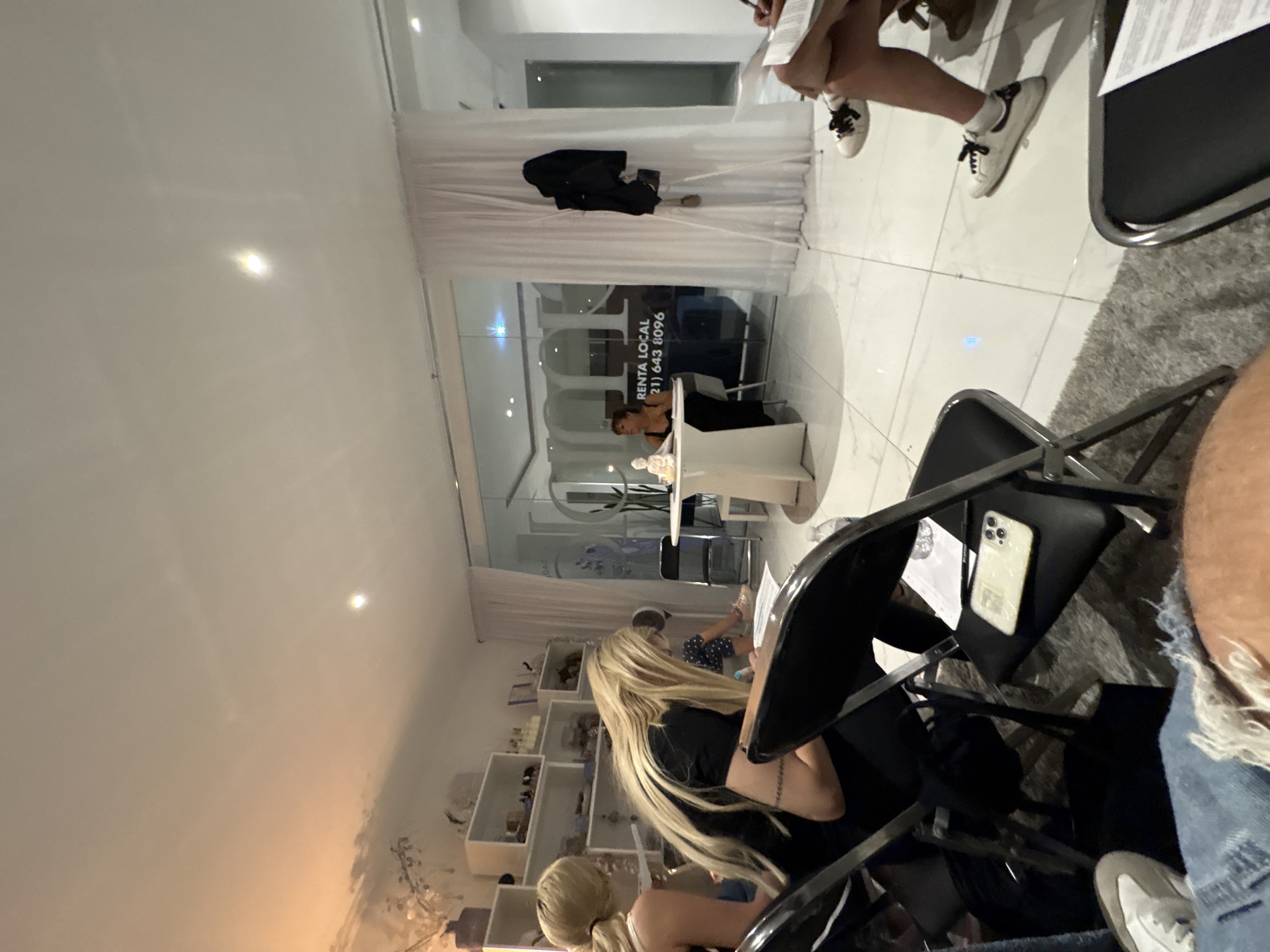 The image shows an interior setting, seemingly a business or event space in Puebla, Mexico, given the partial signage "RENTA LOCAL (21) 643 8096". People are seated in black folding chairs, facing a presenter or focal point beyond the camera's view. One person sits behind a desk. Some individuals are visible in the foreground. The scene is lit with overhead lighting, reflecting off the tiled floor. A white curtain is hung across the back. A decorative bookshelf is placed in the back with assorted items.
