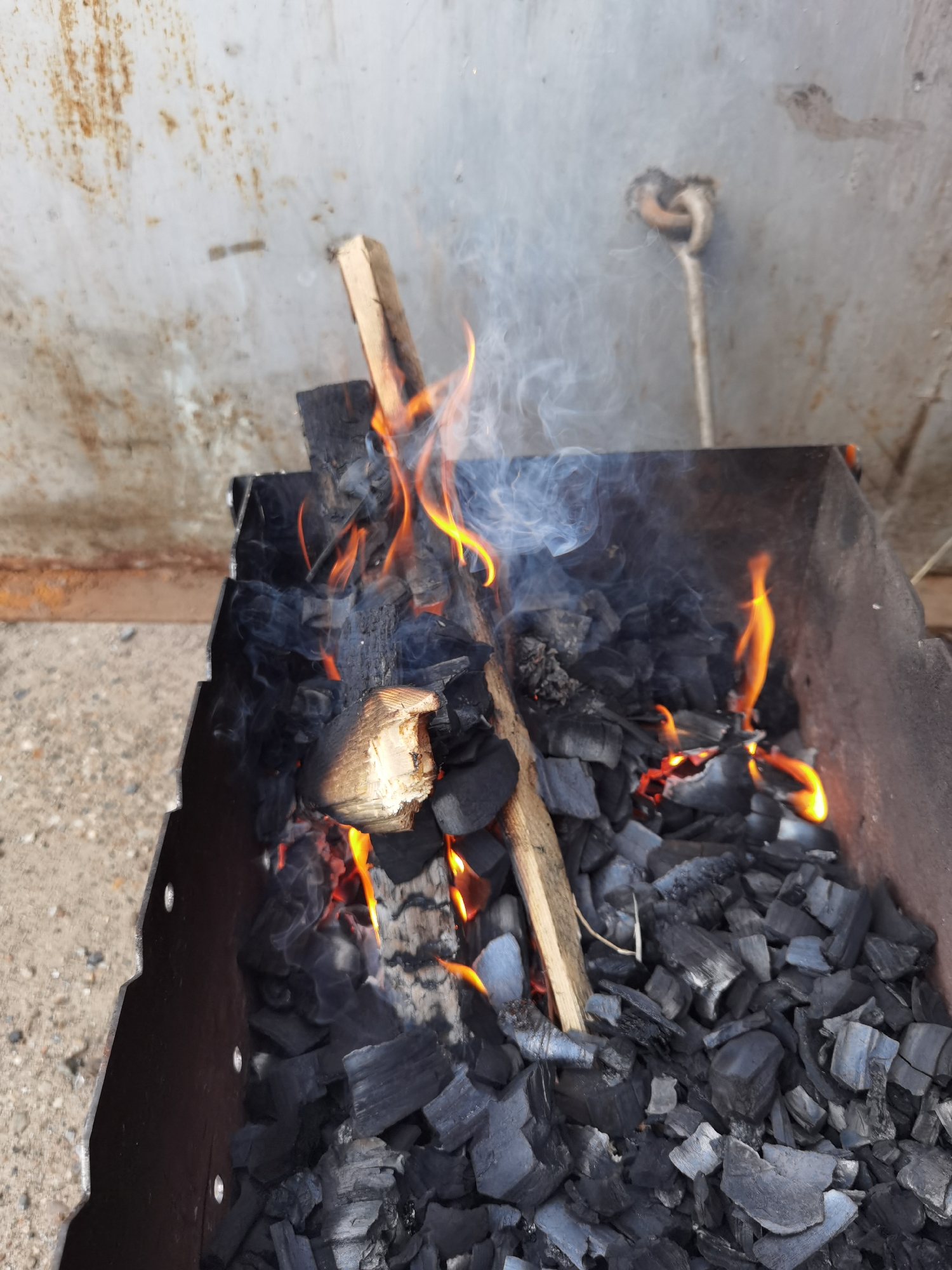 A close-up, slightly high-angle shot captures a metal brazier with burning charcoal and wood kindling, actively producing flames and smoke. The brazier, made of dark metal, appears sturdy with cut-out ventilation along its top edge. Inside, numerous pieces of black charcoal are scattered, interspersed with several pieces of light-colored wood, some of which are actively burning with bright orange and yellow flames. White and grey smoke billows upwards from the burning materials.
The background consists of a light-colored, possibly painted metal or concrete wall, showing noticeable patches of rust, particularly on the left side, and general wear. To the right on this wall, a metallic hook or loop is visible, attached to the surface. The ground beneath the brazier, visible in the lower left corner, is a textured concrete or paved surface. The scene takes place outdoors during the daytime in Chaikovsky, Russia, likely indicating preparation for grilling or a similar outdoor cooking activity. There is no visible text in the image.