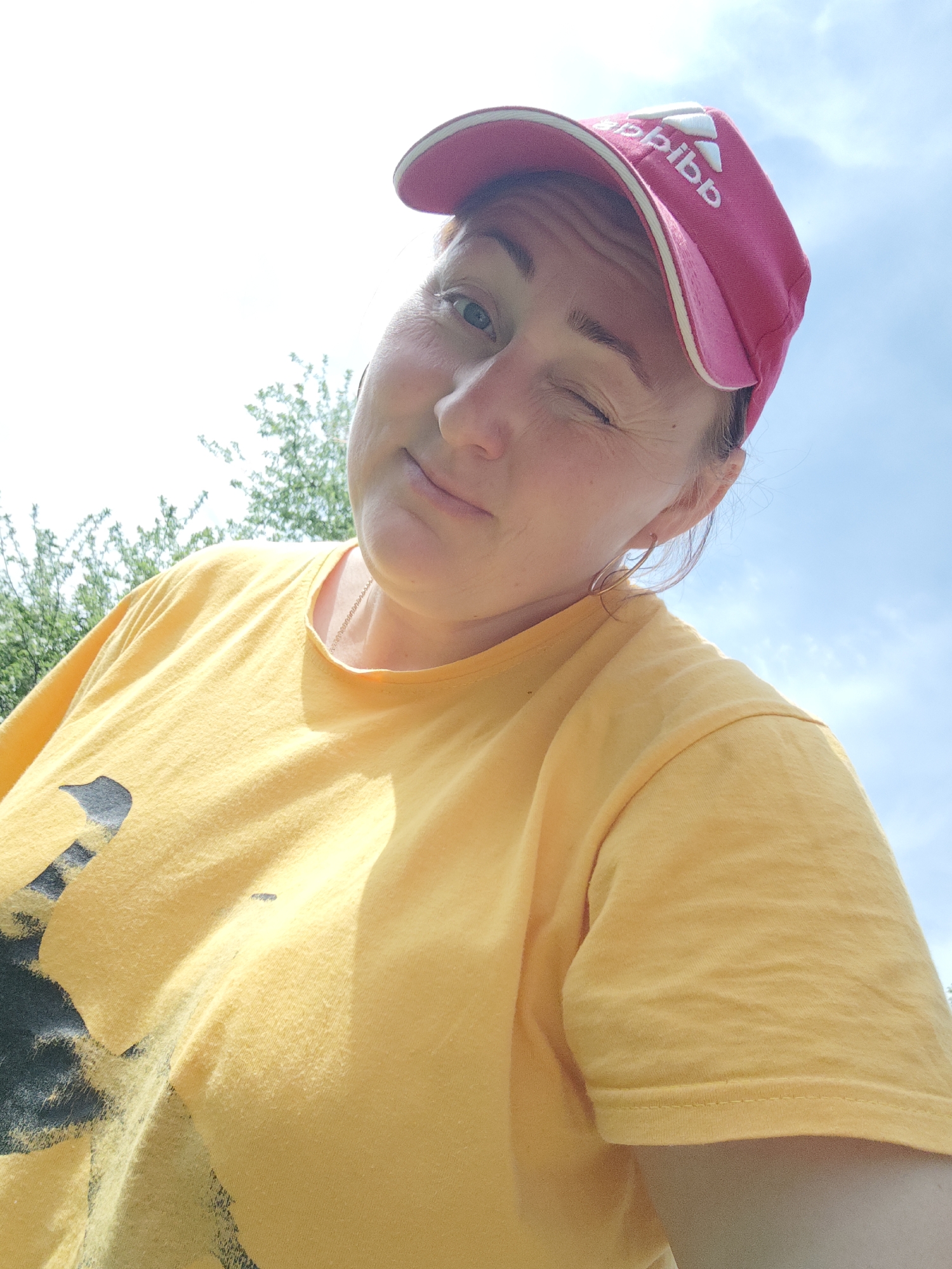 A woman is outdoors, winking and wearing a pink Adidas cap and a yellow t-shirt with a dark-colored graphic on the front. The background includes trees and a blue sky with some clouds. There is no specific evidence to confirm the location as Фоки, Russia.