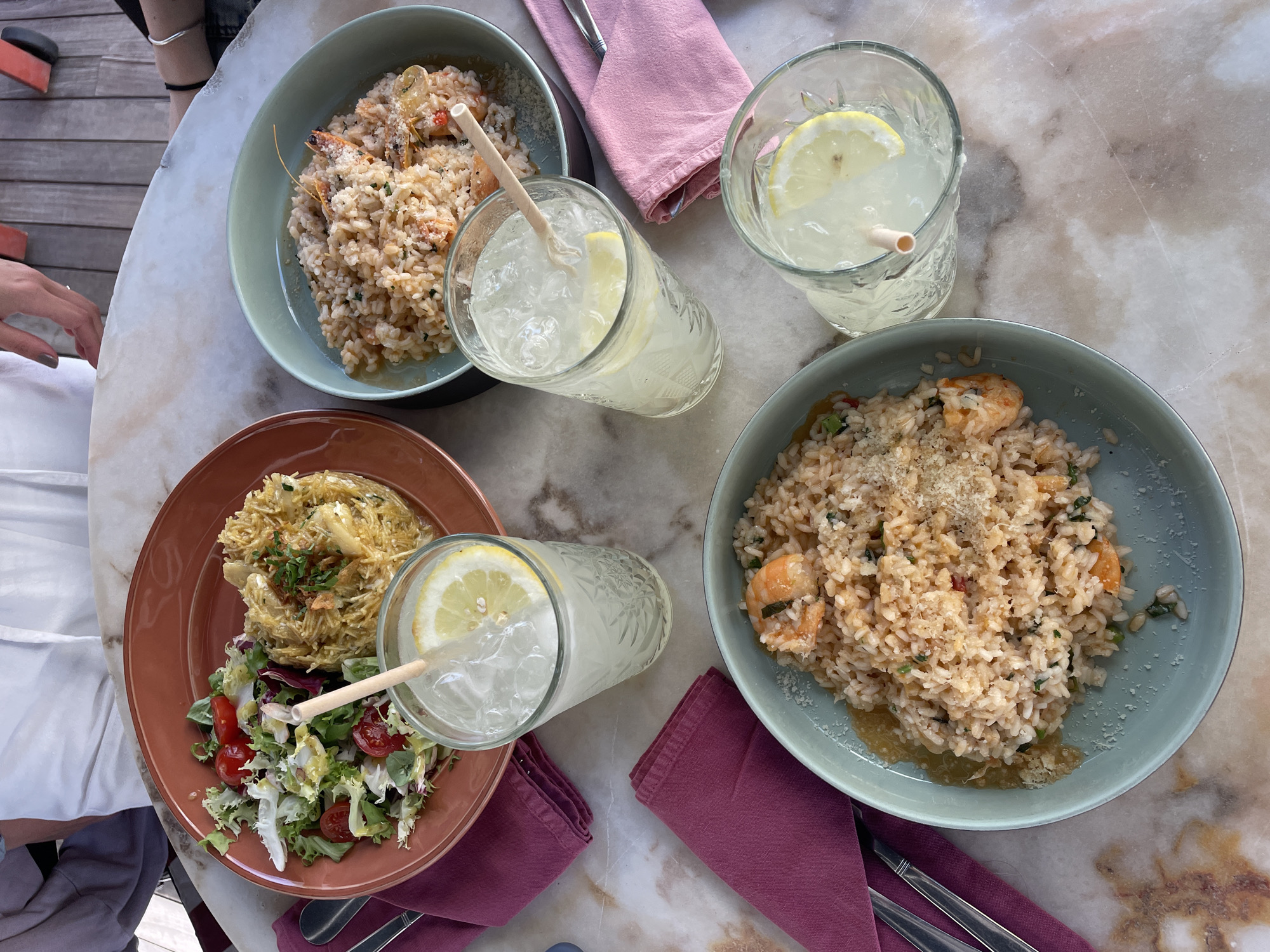 A high-angle shot shows a dining table setting, likely outdoors due to visible wood decking in the background. The table is a light-colored marble with noticeable veining patterns. Four dishes are arranged around the table: two appear to be shrimp risotto in blue-green bowls, one is a salad topped with a pasta-like dish on a reddish-brown plate, and the last looks like a rice dish. Each dish has a corresponding napkin: two are pink, and two are dark red. Three glasses of iced lemonade, each with a lemon slice and a straw, are also placed around the table. Silverware is visible next to one of the dark red napkins. Parts of people are visible on the edges of the photo: hands and clothing, indicating the presence of diners. Based on the dishes, restaurant layout and visible decking, it is implied that this picture was taken outdoors in a dining area, most likely in the city of Porto, Portugal.