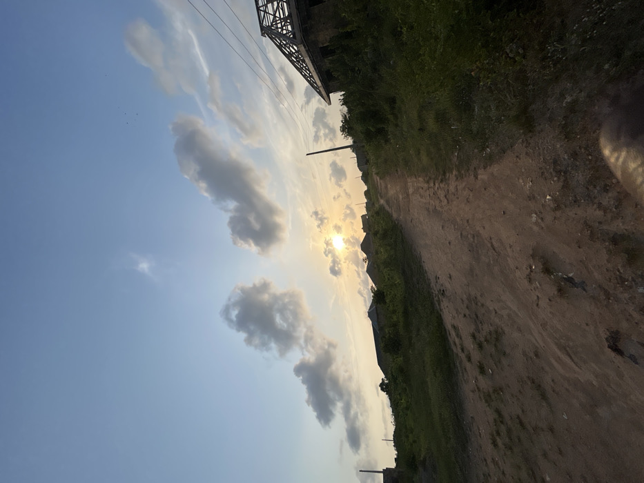The image portrays an unpaved, reddish-brown dirt path ascending a gentle incline in Debasco, Nigeria, during late afternoon or sunset. The sky is light blue with scattered cumulus clouds, and the sun is visible as a bright, partially obscured disc near the horizon, slightly left of center. Dense green vegetation borders the path on both sides. On the right, an unfinished building with exposed concrete block walls and an incomplete wooden roof truss system is prominent, adjacent to a dark utility pole supporting overhead lines. In the mid-ground on the left, several structures with dark roofs and a concrete pillar are partially visible. No individuals, actions, or interactions are discernible. A dark, indistinct object occupies the bottom right corner of the frame.