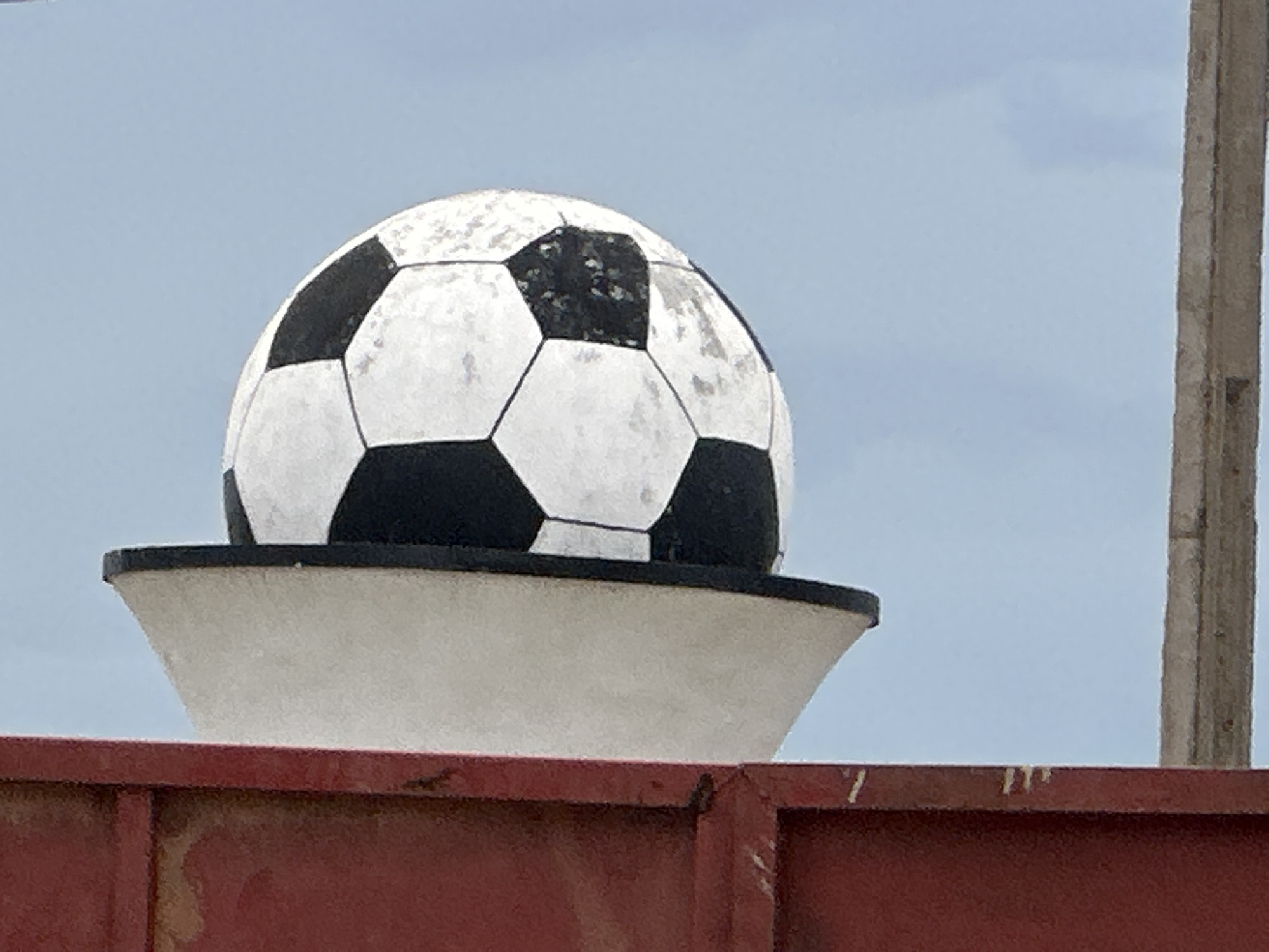 This media file features a close-up, eye-level shot of a large, weathered soccer ball atop a white, curved pedestal. The soccer ball, with its classic black and white hexagonal panels, appears to be a decorative element rather than a functional sports item, given its size and placement. Some of the white panels show signs of wear and tear, with dirt or discoloration.
The soccer ball and pedestal are positioned against a pale blue sky, with a few wispy clouds visible. To the right of the soccer ball, a thick, weathered wooden pole or post is partially visible, suggesting an outdoor setting. In the foreground, the top edge of a reddish-brown metal fence or barrier is present, adding a touch of color and context to the scene. The overall lighting suggests it is daytime, possibly overcast or during a period of diffused sunlight.
There are no people visible in the frame, and no discernible activity is taking place, as the focus is on the static objects. The image conveys a sense of stillness and perhaps a slightly neglected, outdoor environment. The location context of Dadi Riba, Nigeria, could imply a specific cultural or community space where such a decorative soccer ball might be found, perhaps as a landmark or sports-related art installation. No text is visible within the frame.