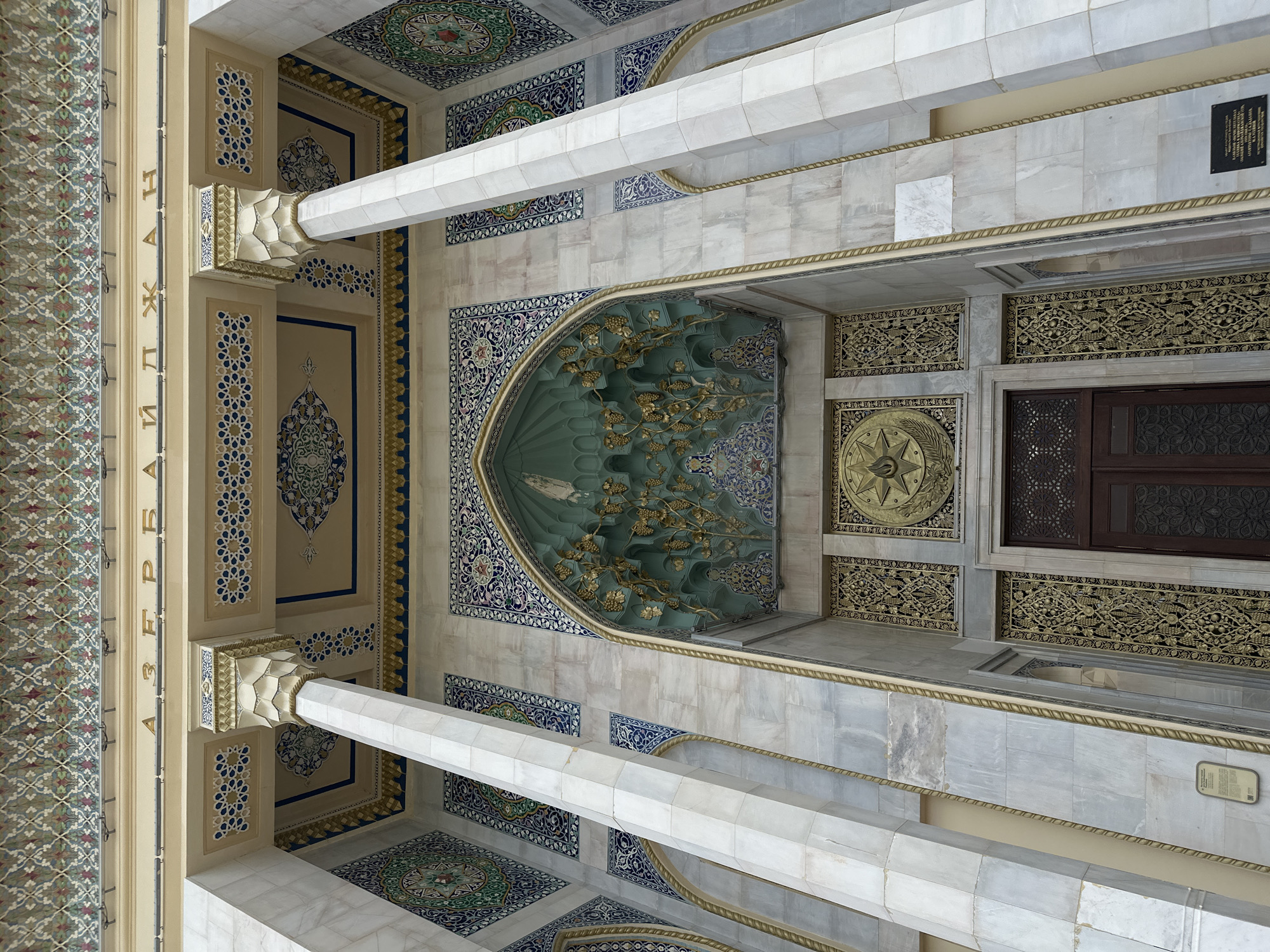 A ceiling view of Heydar Aliyev Mosque in Baku, Azerbaijan showcases intricate Islamic architecture. The walls feature geometric patterns and floral motifs. The color palette primarily consists of white, cream, gold, and blue. The scene captures the interior of a religious building, emphasizing the aesthetic design. The structural columns appear to support the domed roof. The perspective allows for a detailed observation of the craftsmanship.