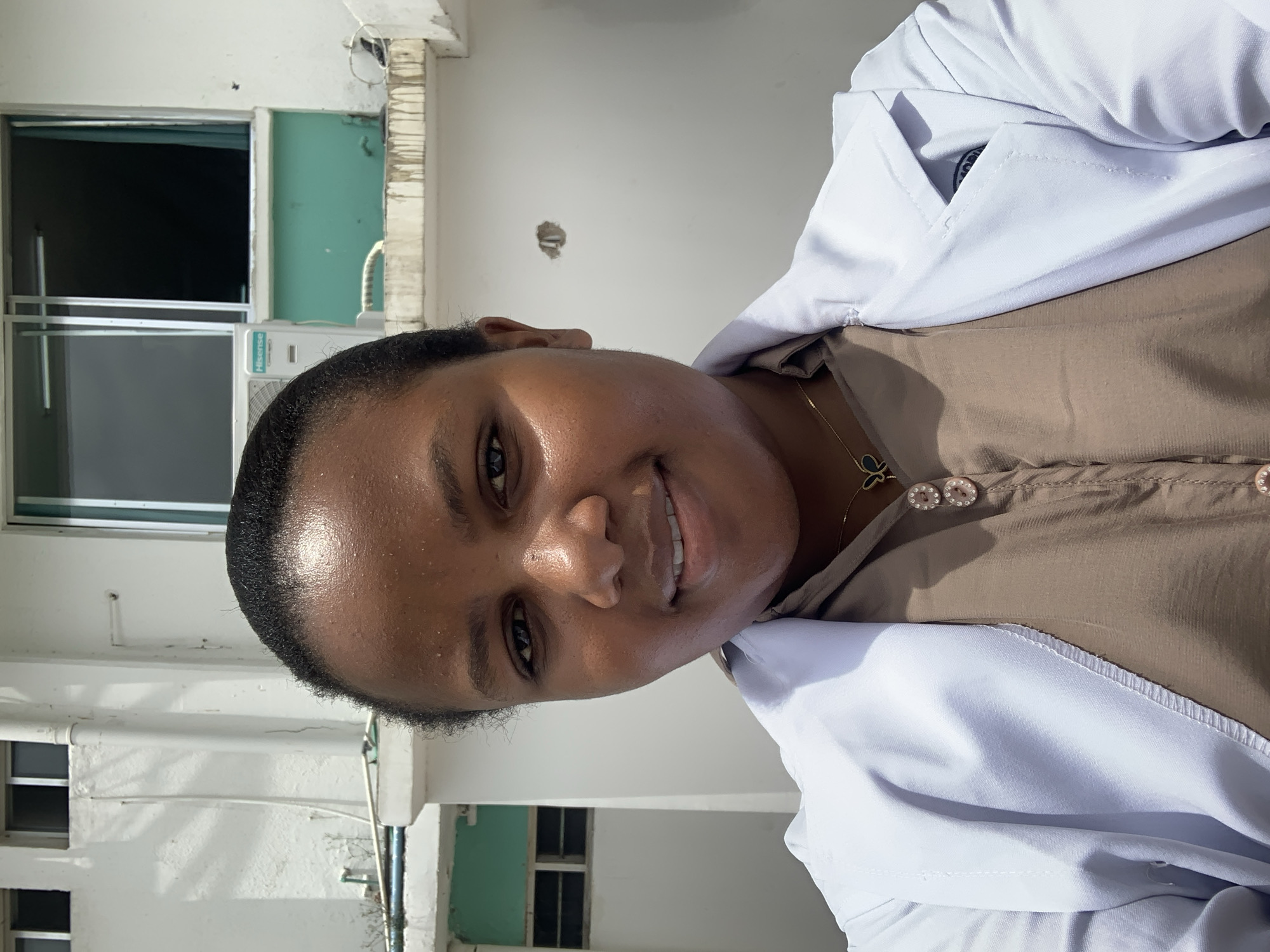 A young adult female is depicted in a close-up outdoor portrait, facing the camera with a faint smile. She has dark, short hair pulled back, and her skin exhibits a visible sheen. She wears a light brown collared shirt fastened with two circular, pinkish-brown buttons, over which she layers a white lab coat or professional jacket. A gold chain necklace with a dark-colored butterfly pendant is visible around her neck.
The background features a light-colored, likely concrete, building, consistent with urban architecture found in Dar es-Salaam, Tanzania. Architectural details include several windows with dark interiors; one large window has a silver-colored frame. Below this window, a section of the wall is painted a distinct teal color, and a white "Hisense" branded air conditioning unit is mounted. A white vertical downspout is visible on the left side of the building. The structure shows signs of weathering, including dark streaks and grime, particularly below ledges and the teal section. Exposed conduits or fixtures are present on the wall to the right. The scene is brightly illuminated by direct sunlight, casting subtle shadows.