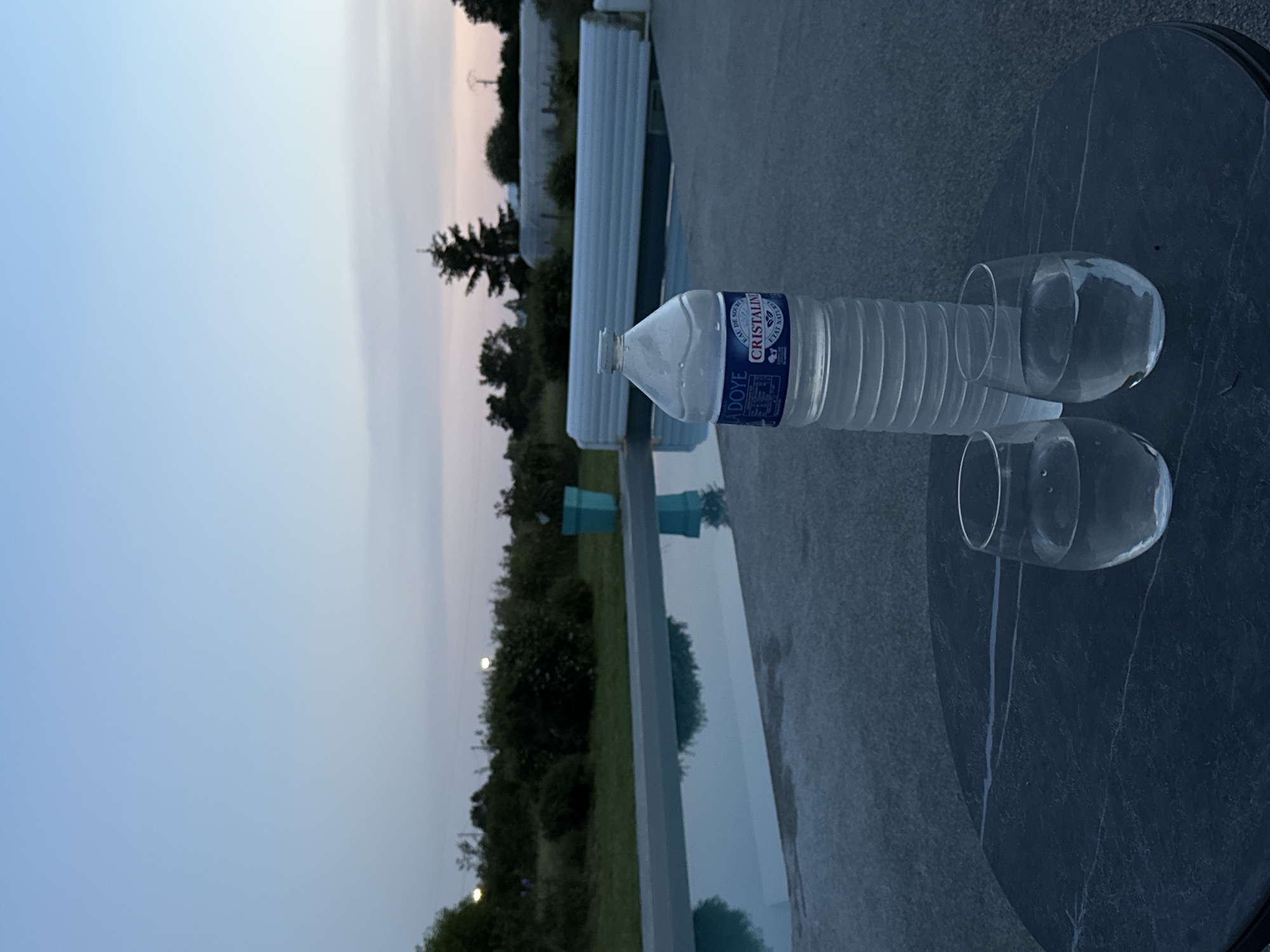 A water bottle and two empty glasses sit on a dark, round, stone-like surface in Veauchette, France. The surface appears to be an outdoor table or countertop next to a swimming pool. The pool's water reflects the surrounding trees and sky, suggesting a location near nature. Beyond the pool and trees, the sky is a gradient of light blues and pinks, indicating either dawn or dusk. The bottle is a "Cristaline" brand water bottle. No people are visible.