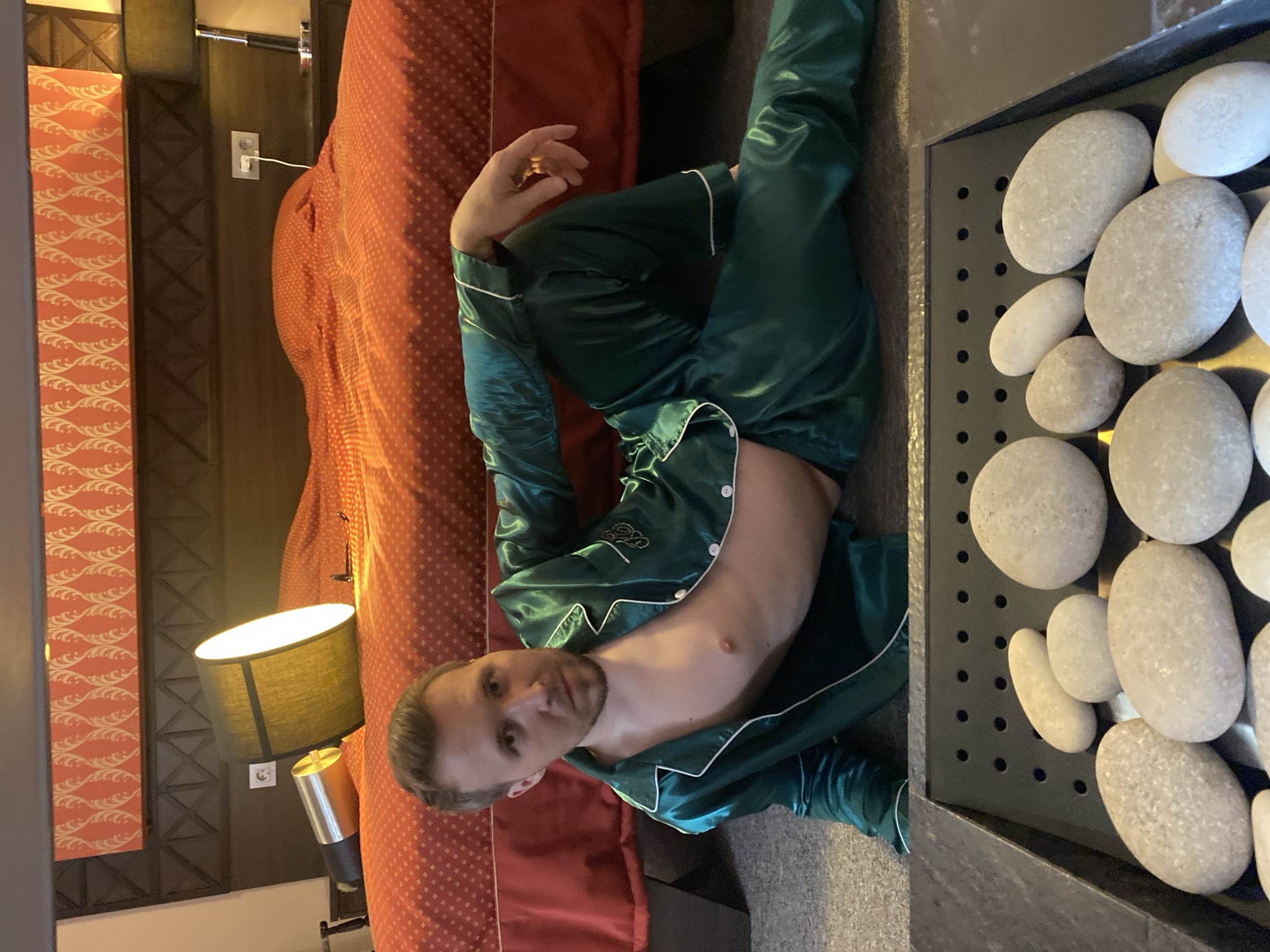 A man wearing emerald green silk pajamas with white piping is lying on a bed covered with a red and white patterned bedspread in a room with beige walls. He is facing the viewer and holding his hand near his head. A black lampshade with a lamp on a pole is on the left side of the image. A black metal rack containing gray stones is on the right side of the image. The image may have been taken in a hotel or home in Yekaterinburg, Russia, but the specific location cannot be determined.