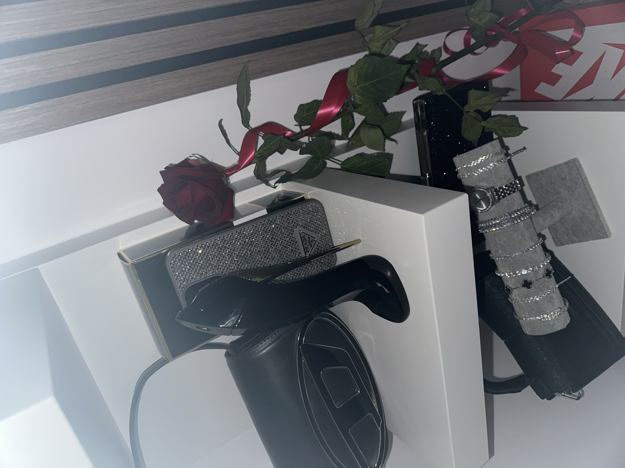 The image shows a collection of luxury items arranged on a white surface, possibly a shelf or tabletop, against a backdrop of white walls and a dark wooden-patterned element (possibly a headboard or part of a furniture piece).
A red rose lies atop a shiny, possibly studded, rectangular object, which might be a clutch or phone case. A pair of black high heels rests on top of a black cylindrical object with a large "D" logo, presumably a purse or accessory by Diesel.
To the right, a black smartphone leans against a grey stand displaying a watch and several bracelets. The setup suggests a curated display of fashionable items. A red ribbon wraps around the stem of the rose and extends towards a red and white box in the upper right corner, adding a touch of elegance.
The location appears to be indoors, possibly a bedroom or dressing area in an unknown city. There is no presence of people visible in the frame.