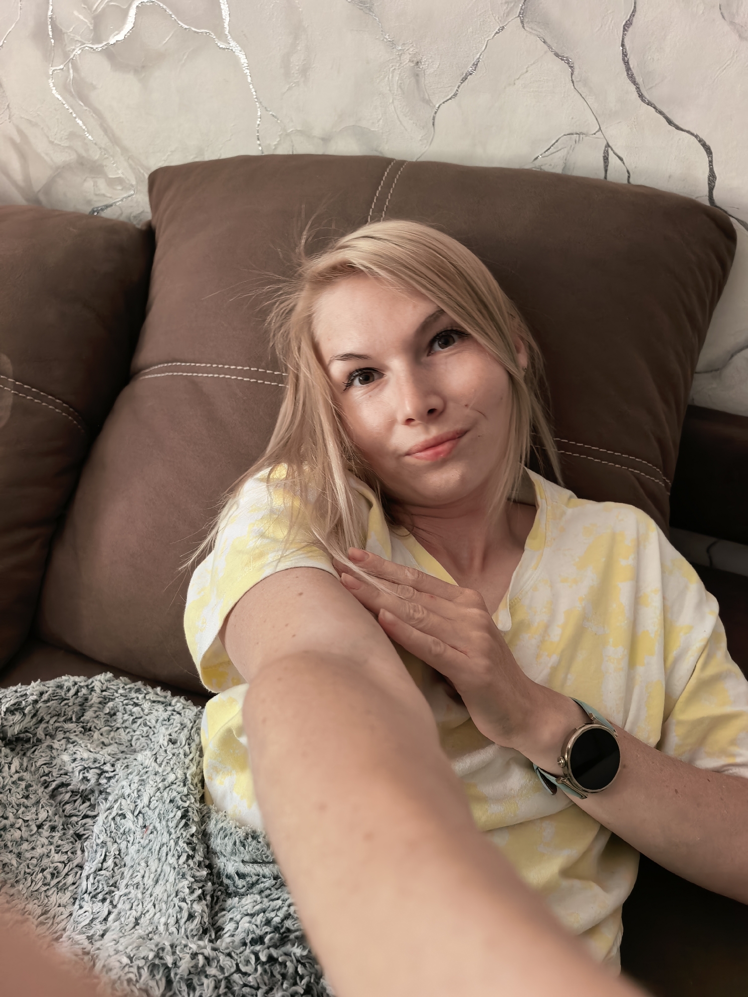 A fair-skinned woman is lying down on a brown sofa with a gray-blue blanket, taking a selfie. She is wearing a yellow and white tie-dye t-shirt and a smartwatch with a light-colored band. The wall behind the sofa has a textured, marble-like pattern with silver accents. The location is likely a private residence. There are no explicit indicators that the location is in Sterlitamak, Russia.