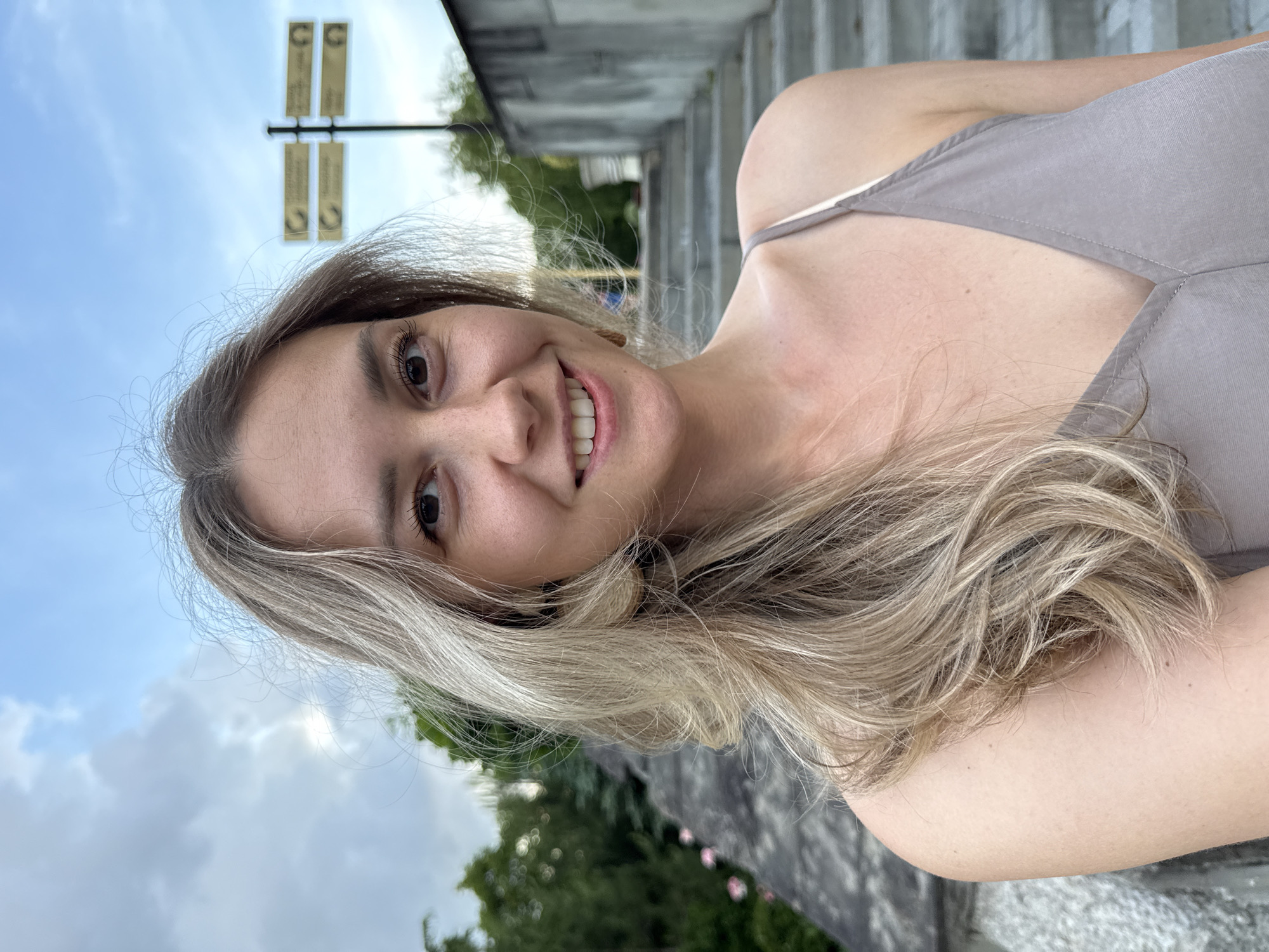 A woman with blonde hair smiles into the camera, holding it at arm's length to take a selfie. She is outdoors, with a grey stone wall and stairs behind her. Green trees and a blue sky with white clouds are visible in the background. There are two street signs on a pole behind her. The woman is wearing a light brown camisole. The location is Nebug, Russia.