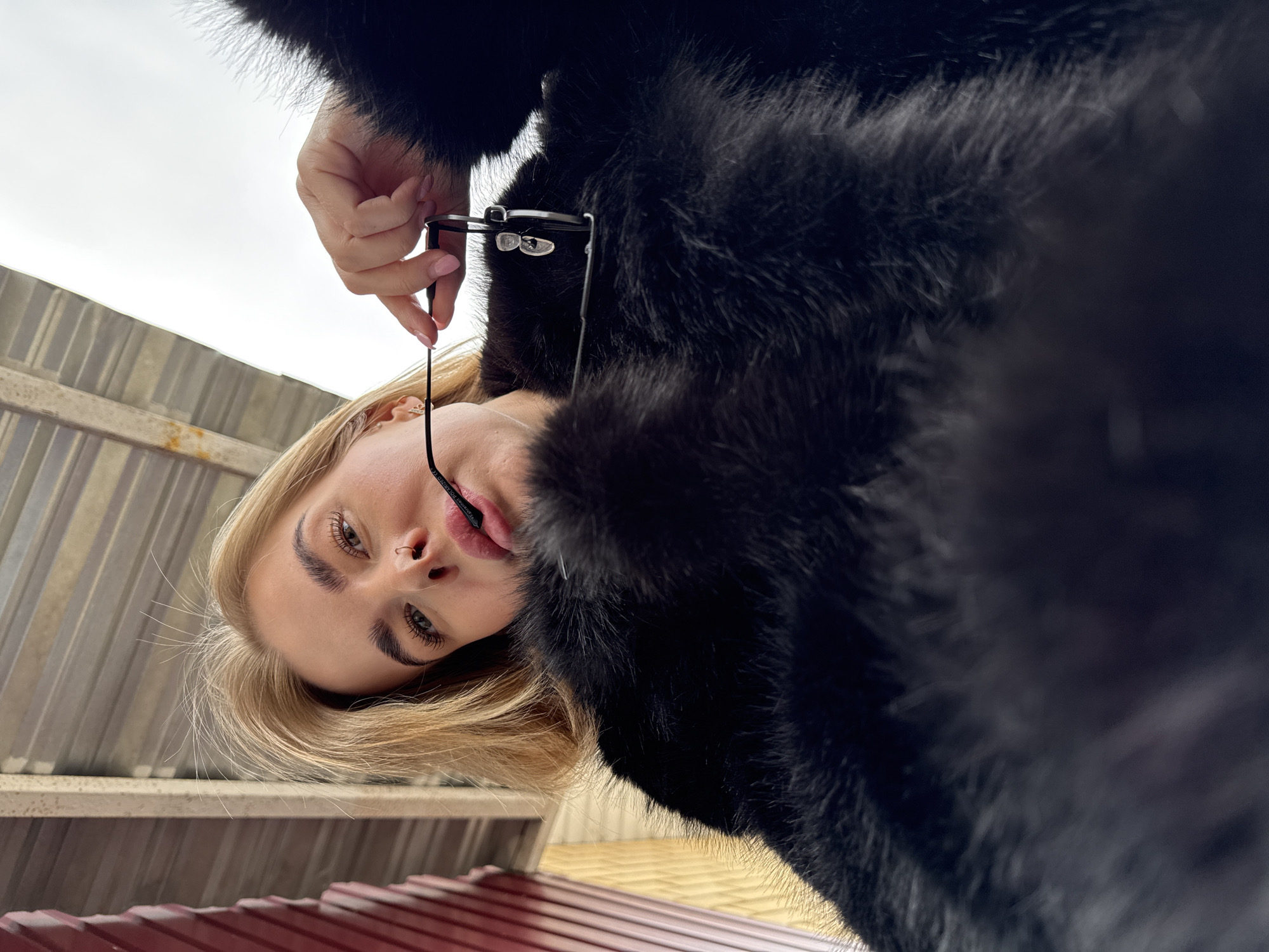 A low-angle shot features a blonde woman wearing a black fur coat and holding a pair of glasses, taken in Chaikovsky, Russia. The view is from below, looking up at the woman who is tilting her head. Her blonde hair is styled and her facial features, including her eyes, nose, and lips, are clearly visible. She has a nose ring. She is holding the eyeglasses by the frame with her right hand. The background consists of a metallic, slatted surface in muted tones of silver and red. The sky is overcast.