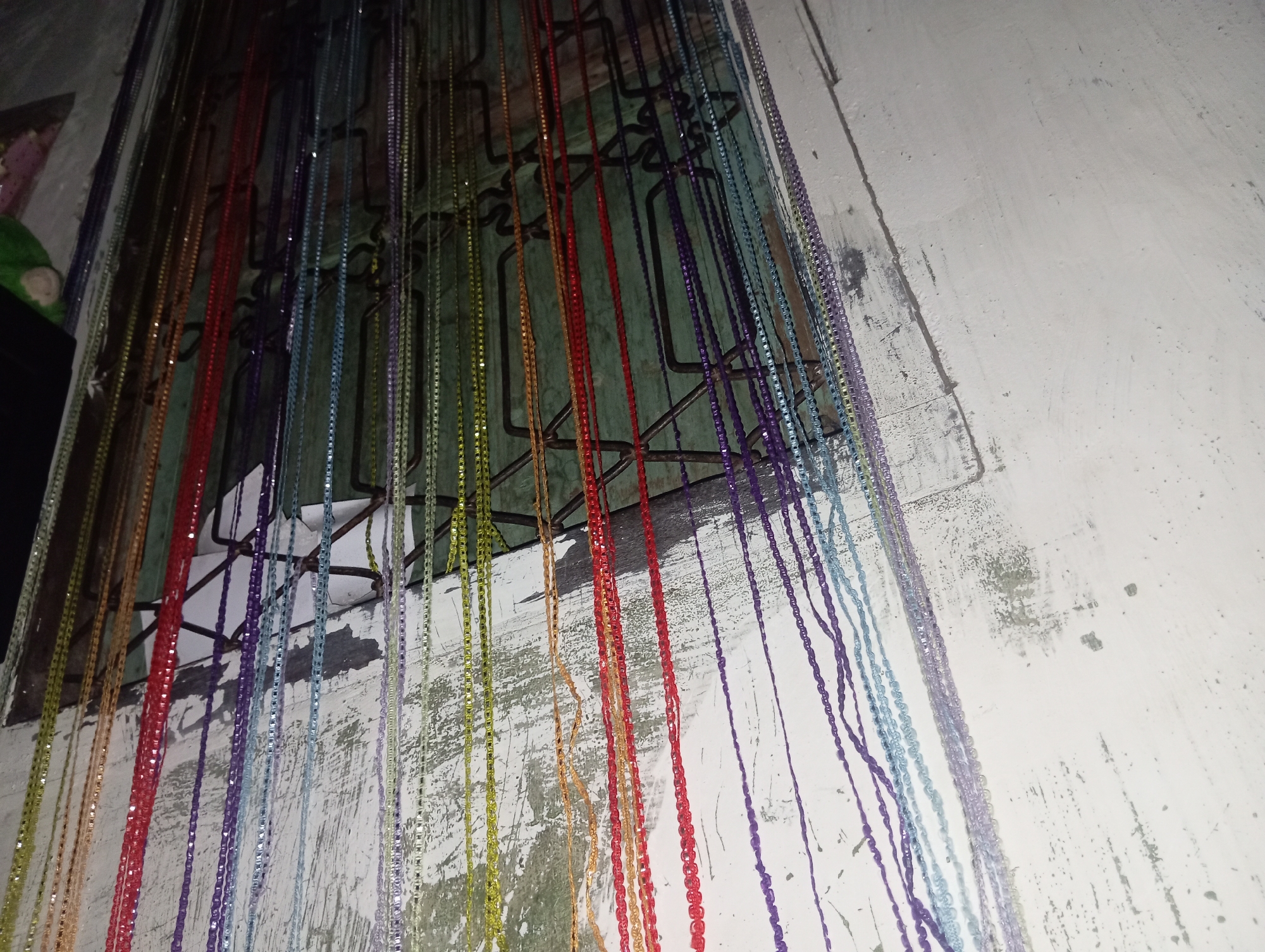 A beaded curtain hangs in front of a green-painted window with a black metal grate. The curtain is made of numerous strands of small, colorful beads in shades of purple, blue, light blue, red, orange, yellow, and green. The beads are strung together to form a dense curtain. Behind the curtain, the window frame appears to be made of dark wood, and the grate is made of twisted metal bars. To the right of the window is a white wall with peeling paint. The wall has visible cracks and patches of exposed material. To the left of the window, a dark object, possibly furniture, is partially visible, with a glimpse of a pink fabric and a green object, possibly a stuffed toy, on top of it. The lighting in the image is dim, suggesting an indoor setting. The overall scene depicts a cluttered entryway or room in what appears to be a residential area in Rejosari, Indonesia.