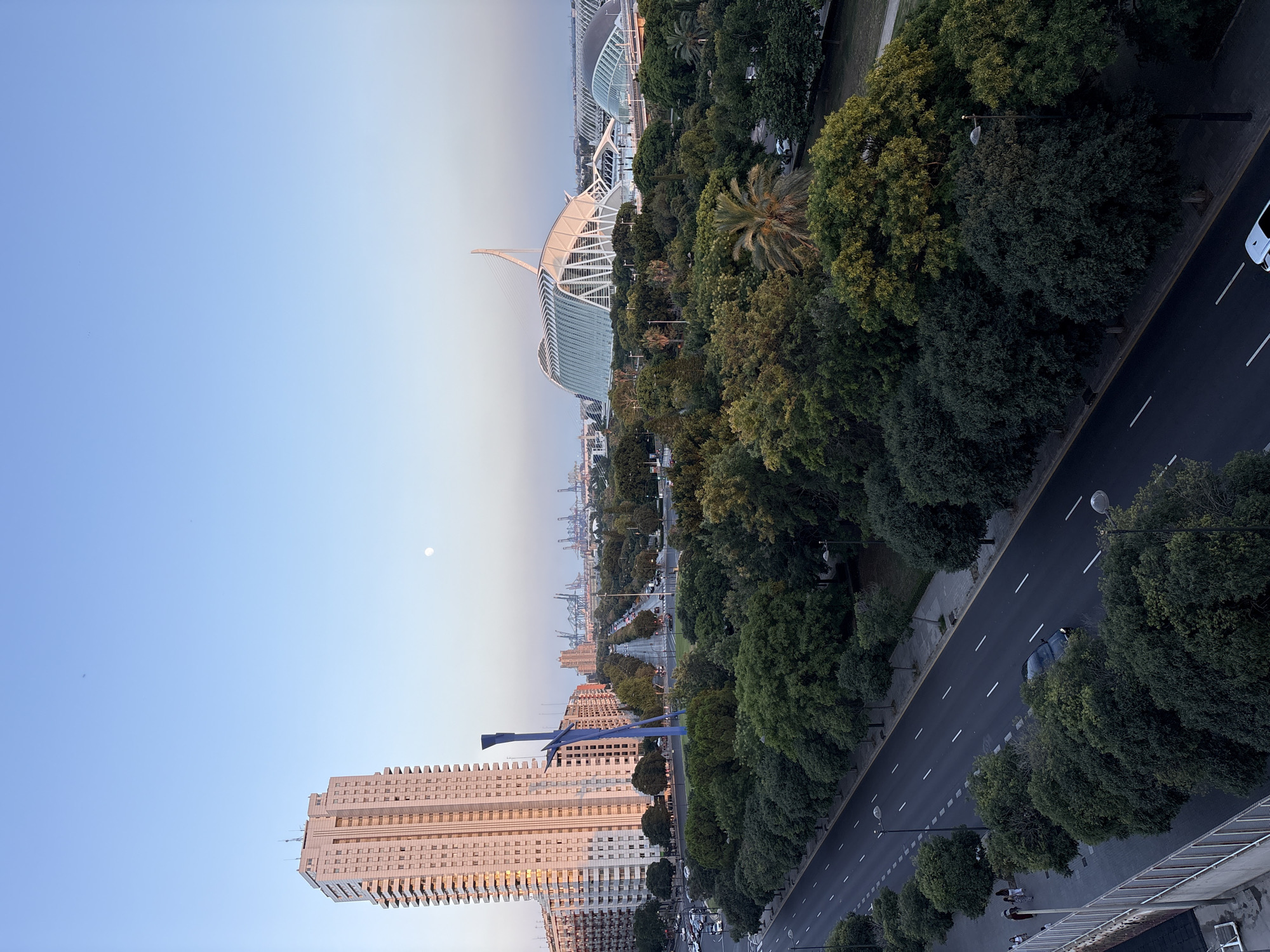 This is a high-angle, wide-shot photograph of the City of Arts and Sciences in Valencia, Spain, taken during the golden hour. In the foreground, a multi-lane road with a lone car driving on it runs parallel to a tree-lined sidewalk. A light-colored building with many windows and balconies dominates the left side of the frame, with a tall, blue, abstract sculpture standing in front of it. The middle ground is a lush green park filled with trees. In the background, the distinctive white, futuristic architecture of the City of Arts and Sciences, including the Hemisfèric, is visible. The sky is a clear, light blue, with a faint moon visible. The overall scene depicts a serene urban landscape as the day draws to a close.