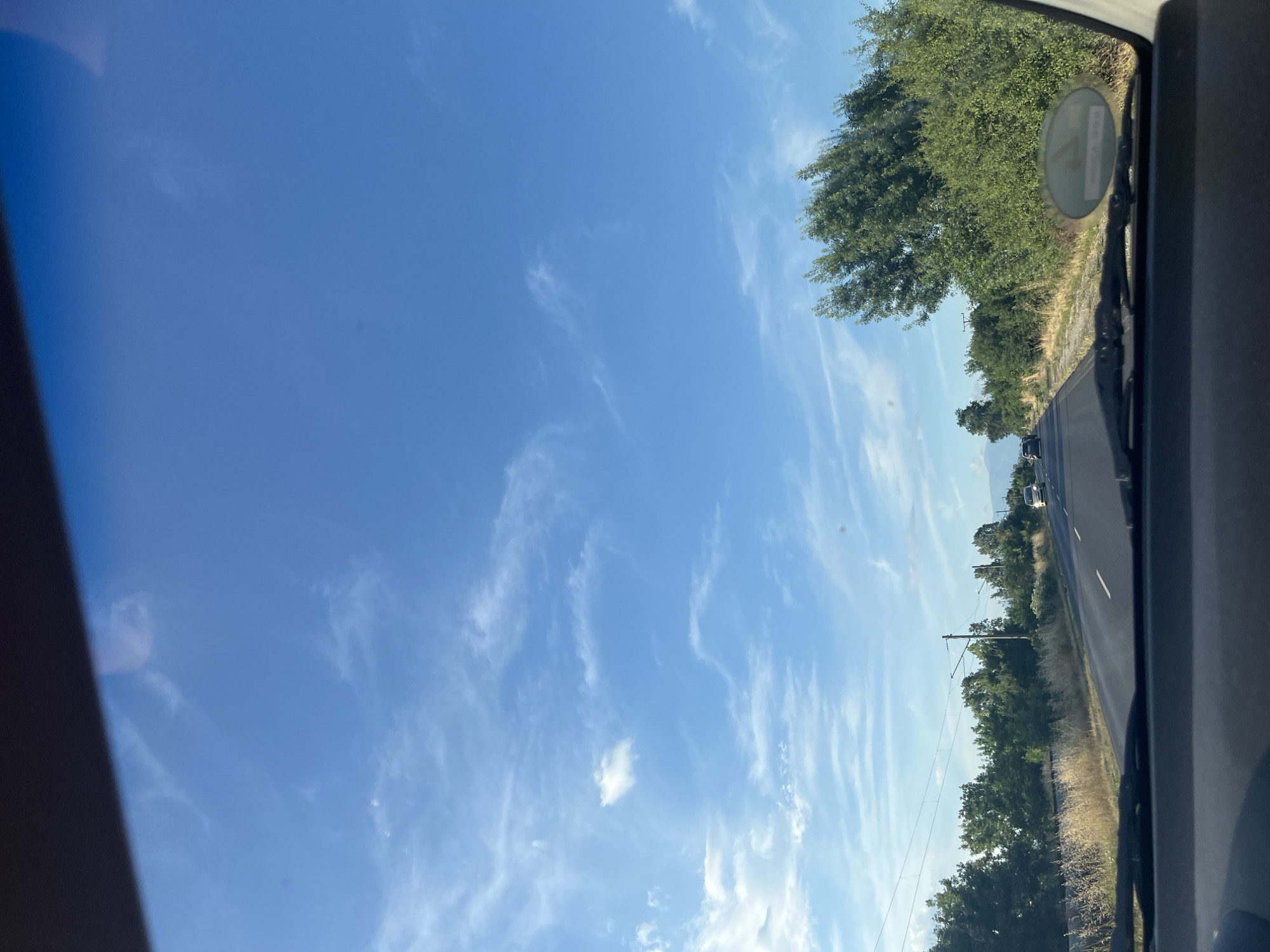 The image shows a view from inside a vehicle looking out through the windshield onto a road. The road is asphalt and appears to be a highway with at least two lanes in the direction of travel. There are cars visible on the road, moving in the same direction. The surrounding environment consists of a grassy shoulder with dry vegetation, followed by a line of trees and bushes. Overhead, there are power lines and poles. The sky is a clear blue with wispy white clouds. The lighting suggests it is daytime, likely late afternoon or early evening. The perspective is that of a passenger or driver, looking forward. Based on the provided context, this scene is located in Dalgo Pole, Bulgaria.