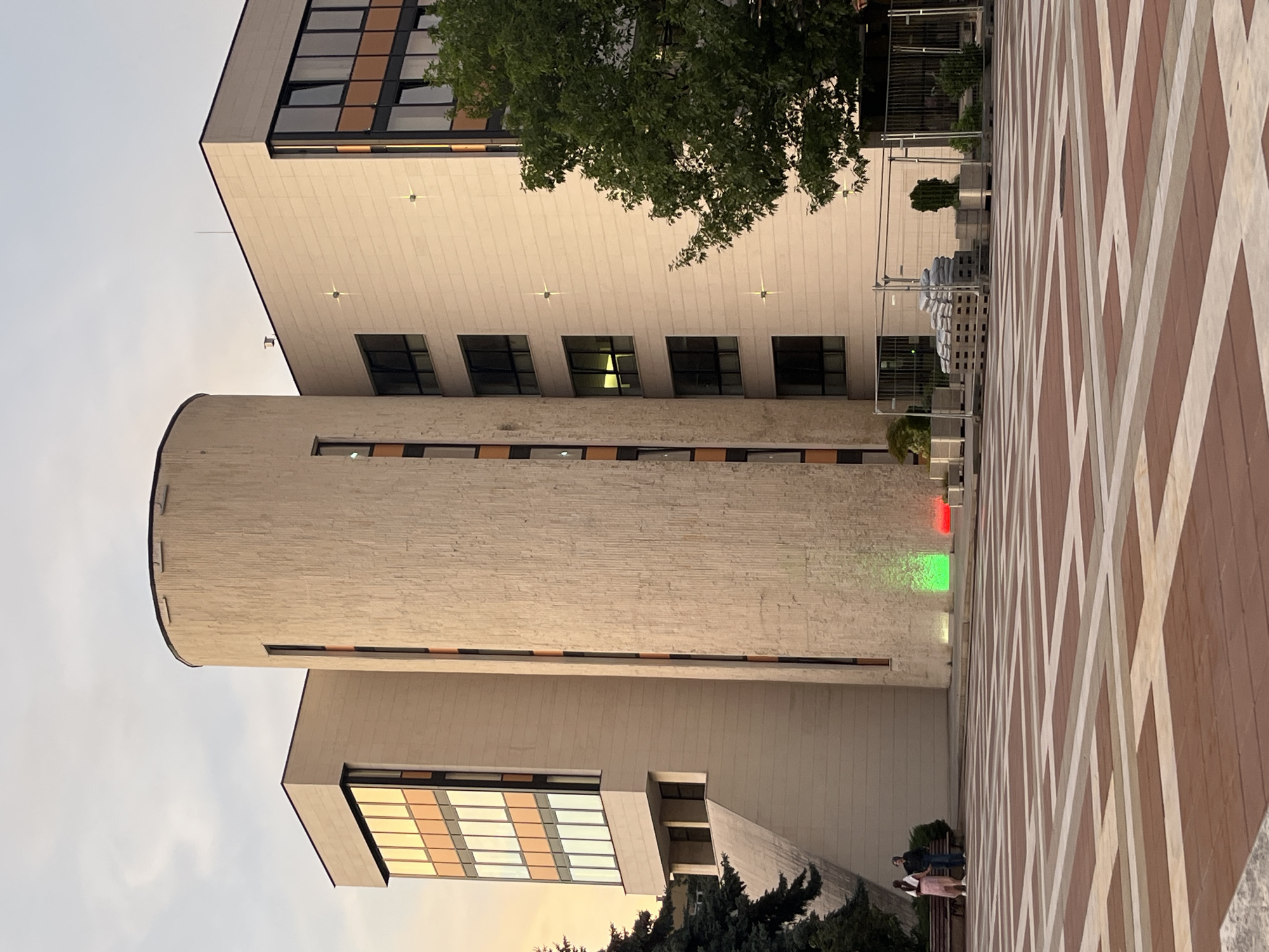 The image captures a section of a modern architectural complex under a pale sky, possibly at dusk, with warm light reflecting off the building's facade. The main structure is characterized by a tall, cylindrical tower with a rough, textured exterior, juxtaposed with a rectangular building section featuring large windows with orange-tinted panes. The ground level is paved with a distinctive geometric pattern of red-brown and cream-colored tiles. In the foreground, two individuals, one in a pink dress and the other in a dark jacket and jeans, are standing near a bench. To the right, a section of the plaza is cordoned off with metal fencing, enclosing stacks of sandbags on pallets and potted plants. A large, mature tree with dark green foliage stands to the right of the building. At the base of the cylindrical tower, two colored lights cast green and red glows onto the pavement. The scene is tranquil, with no overt signs of activity beyond the static presence of the people and the fenced-off area.
