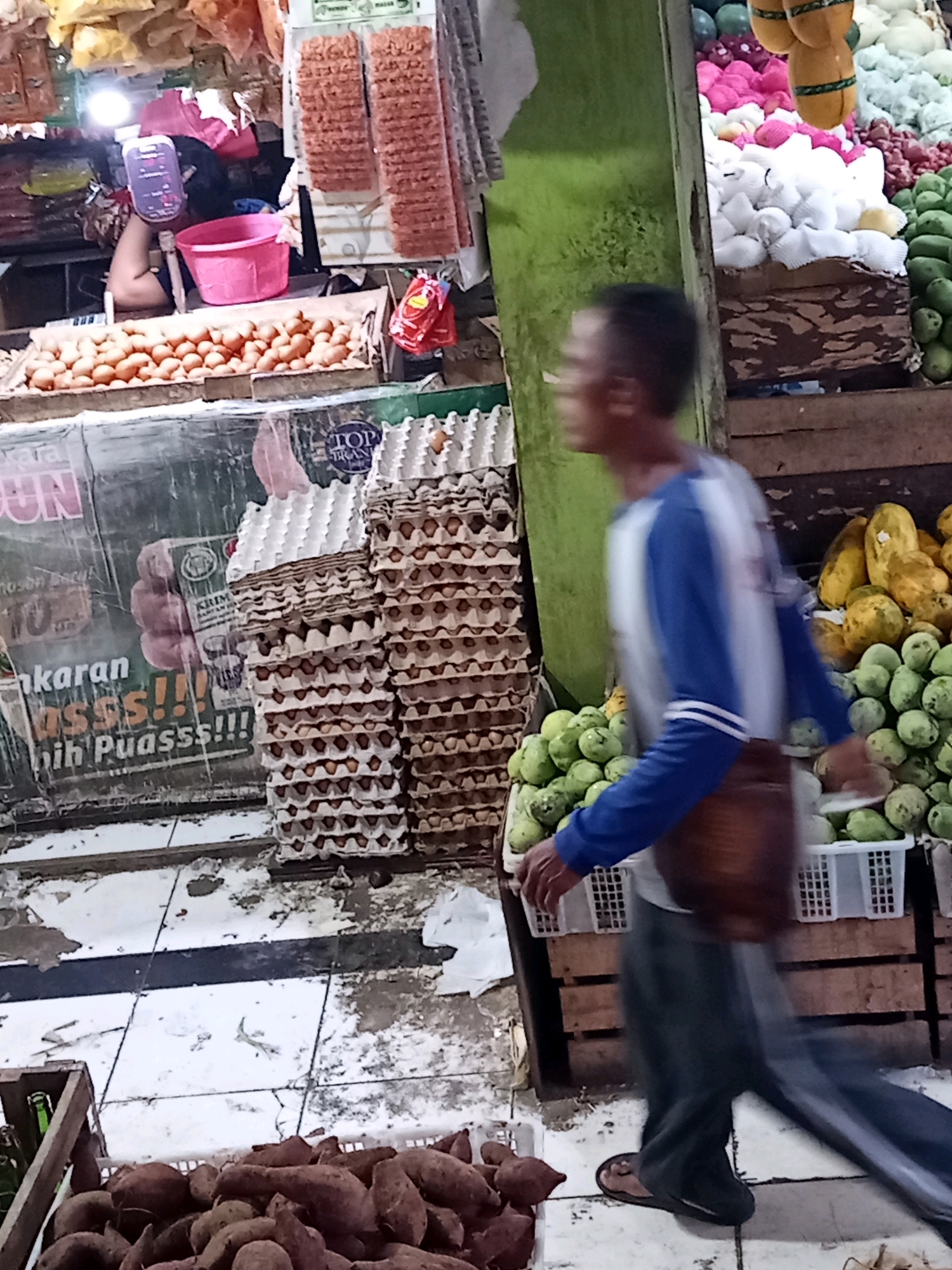 The image captures a bustling market scene in Karawang, Indonesia, likely in a wet market or a street-side stall. In the foreground, a man is walking away from the camera, his body blurred by motion. He is wearing a blue and white long-sleeved shirt, dark pants, and flip-flops. A brown bag is slung over his shoulder. He appears to be navigating a cluttered environment.
To the left of the man, stacks of egg cartons filled with eggs are prominently displayed, suggesting a food stall. Behind the egg cartons, a large banner with Indonesian text and a graphic of a hand giving a thumbs-up is visible. Further back, a woman is partially visible behind a counter, tending to a pink bucket and a pink plastic container. Shelves above are stocked with various items, including what appear to be snacks or packaged goods.
To the right of the man and behind him, a colorful assortment of fruits is arranged in crates and baskets. There are yellow papayas and green fruits, possibly mangoes or other local produce. The market floor is tiled and appears somewhat worn and dirty, with scattered debris. The overall impression is of a busy, active marketplace filled with produce and goods for sale.