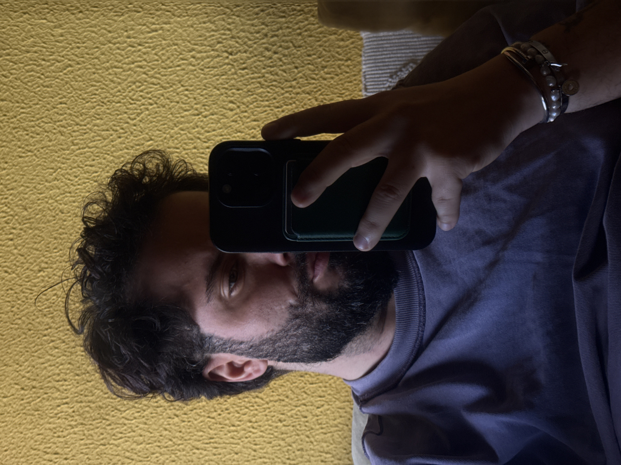 The image displays an adult male, with dark, curly hair and a dark beard and mustache, situated indoors. He wears a light purple, long-sleeved crew-neck shirt. The man's left hand holds a black smartphone, which features a dark green accessory attached to its rear and is oriented towards his face, obscuring part of his left side. His right eye is visible, directed towards the phone. The left wrist is adorned with multiple bracelets, including silver-toned bangles and a strand of pearls. The background is a textured, mustard-yellow wall. A beige striped fabric object, consistent with a cushion or part of a couch, is partially visible on the right side behind the subject. The scene is identified as being within the city of Vila Nova de Gaia, Portugal.