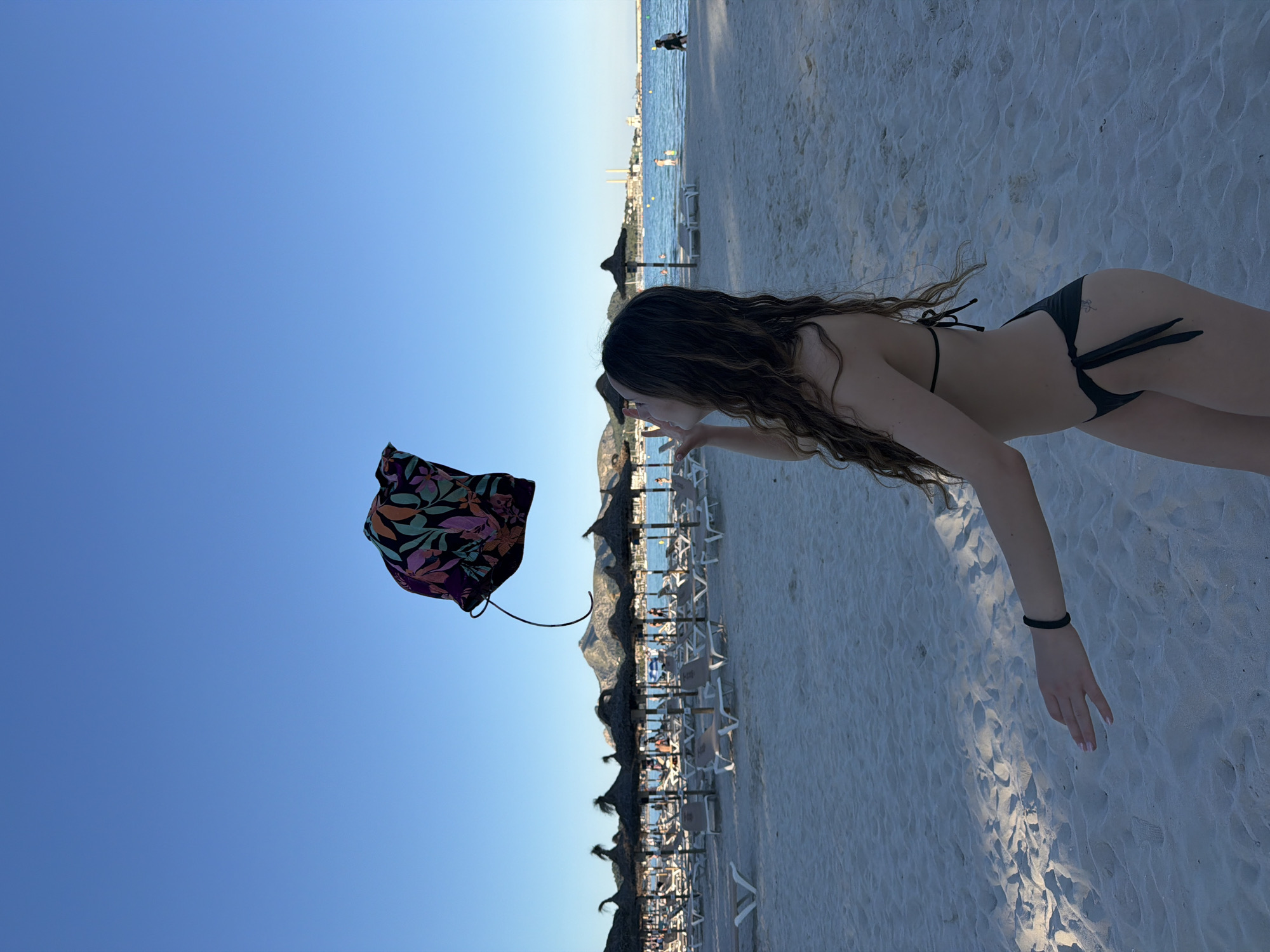 A horizontal outdoor shot, likely taken at Platja D'alcúdia, Spain, features a woman on a sandy beach under a clear blue sky. The scene is arranged so that the sky is above and the beach is below. The woman is wearing a dark-colored bikini and has long, wavy hair. She is leaning forward, with her right arm extended towards the beach and her left arm reaching towards a floral-patterned object floating in the air. Behind her, the beach is lined with rows of beach umbrellas and lounge chairs. Further in the background, the sea meets the horizon, with some buildings visible on the coastline. The sand appears white and textured. The image is well-lit, suggesting it was taken during daylight hours.