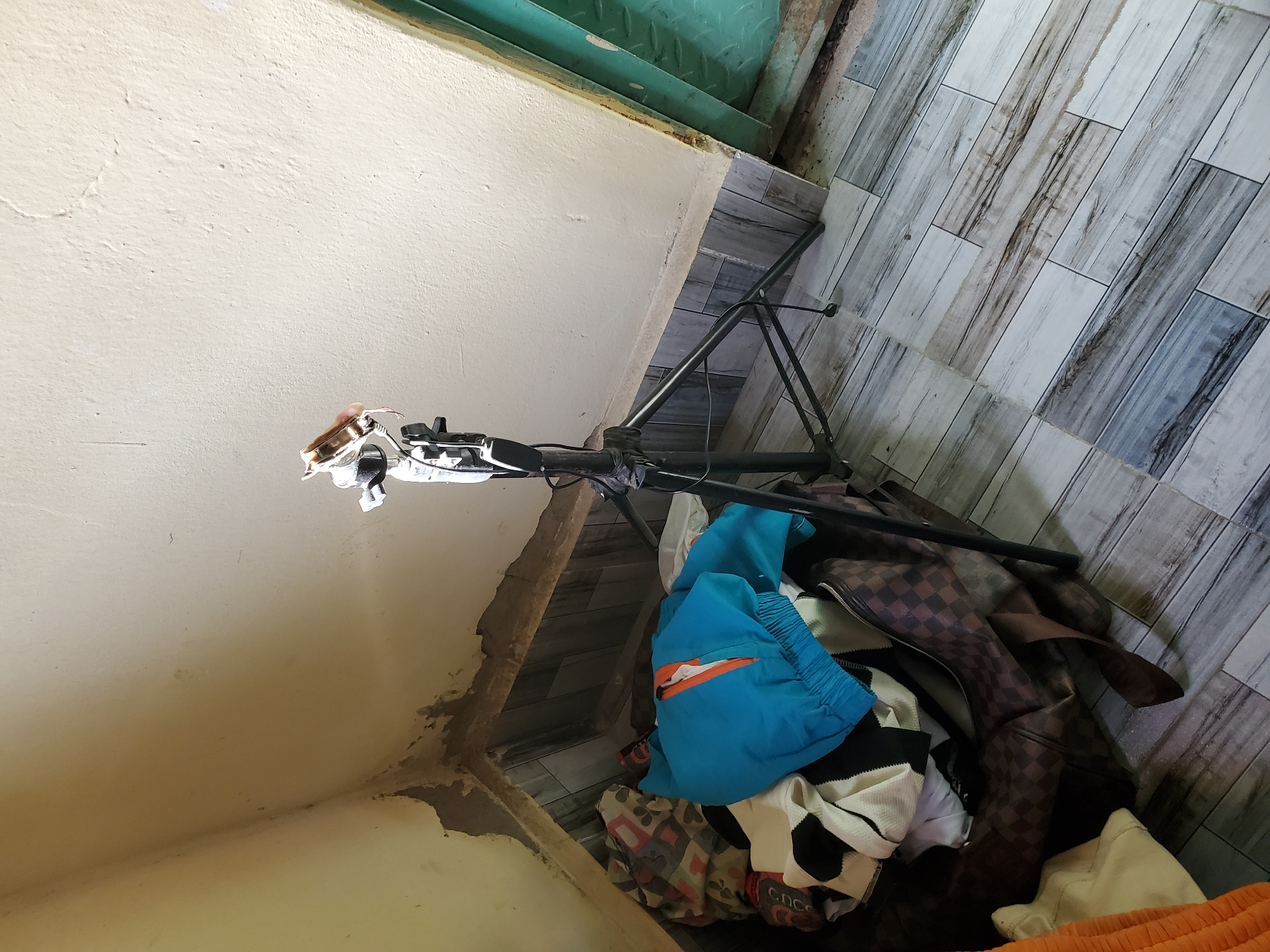 The image depicts a cluttered corner of a room, likely in Jalingo, Nigeria. In the foreground, a collection of clothing and a large, checkered brown bag are piled on a floor tiled with a wood-grain pattern. A blue jacket with orange accents is prominently displayed atop the pile. To the right of the clothing, a black tripod stands, holding a small device at its apex. The background features a cream-colored wall with visible peeling paint and water damage near the floor. To the far right, a green metal door is partially visible. The lighting suggests it is daytime, with light streaming from an unseen source, casting shadows.