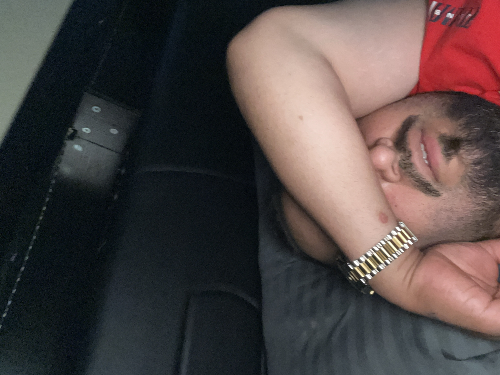 A close-up, dimly lit image shows a man resting or sleeping, oriented with his head towards the right side of the frame. He is seen from the chest up, with his left arm bent and partially covering his face, though his right eye, nose, and mouth are visible. He has dark hair or stubble on his chin and a mustache. His mouth is slightly open, showing some teeth. He wears a red shirt, with some white, unclear text visible on the shoulder area (possibly starting with "SNI"). A two-tone watch with alternating gold and silver links is prominent on his left wrist, which is resting near his face.
The man is lying on a dark, possibly black or very dark grey, upholstered surface, which could be a bed, couch, or vehicle seat. The background to the left is also dark and out of focus, revealing what appears to be a rectangular panel with four small, glowing white circles or lights, possibly indicators or buttons on a control unit, though its exact nature is unclear due to blurriness and dimness. The overall lighting suggests an indoor setting, possibly at night or in a low-light environment. The man appears peaceful in his resting state.