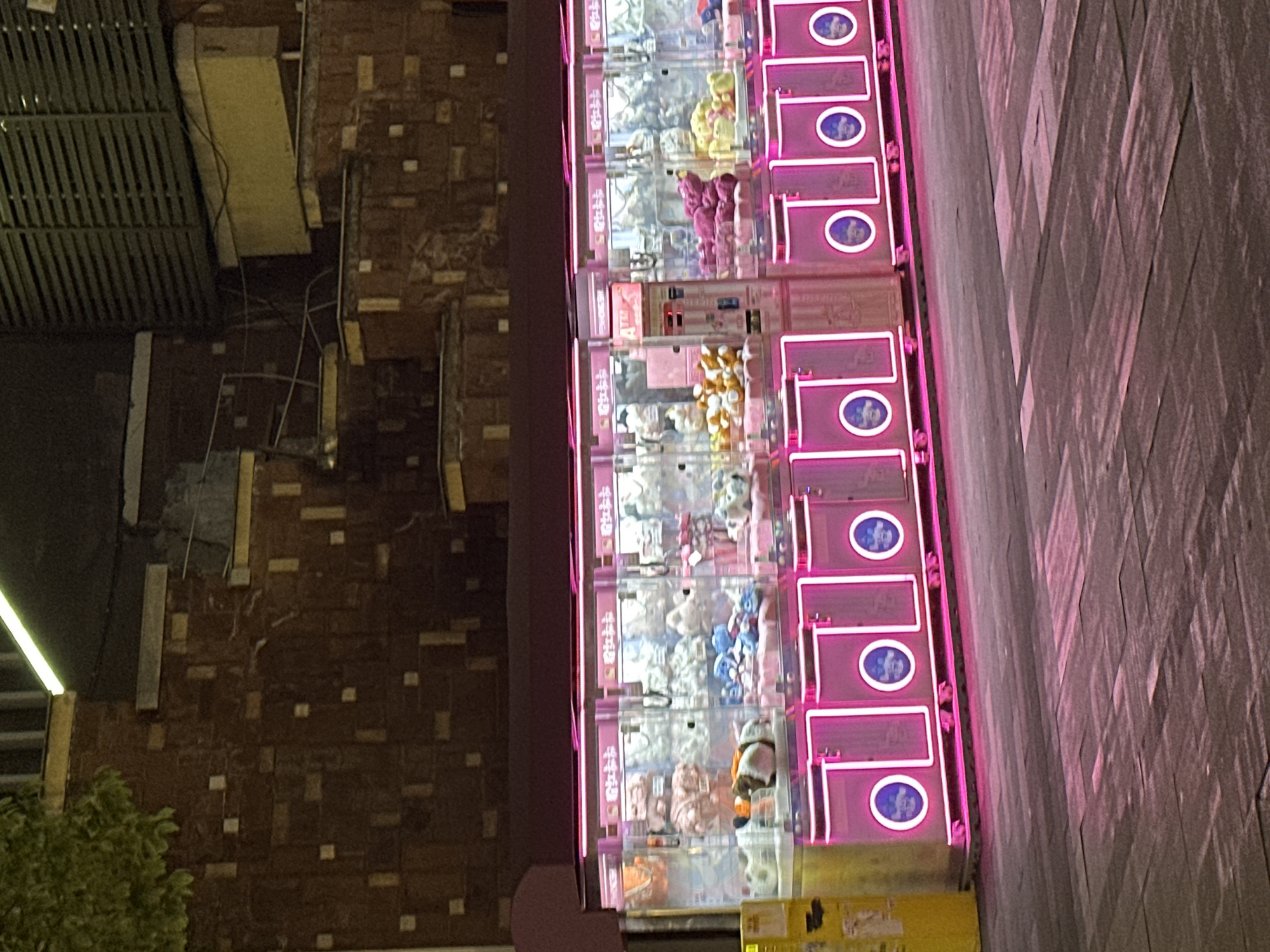 In the city of Chongqing High-tech Zone, China, a row of five claw machine arcade games is brightly illuminated by pink neon lights. The games are filled with an assortment of plush toys, including teddy bears, animals, and cartoon characters. The machines have glass fronts and metal bodies with coin slots and controls. In front of the machines, the ground is paved with interlocking stones that reflect the neon glow. The background consists of a brick wall and some industrial-looking structures. A portion of a yellow vending machine is visible to the far left of the image. There are no people or animals present in the scene.