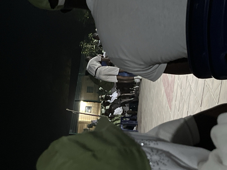 An outdoor gathering at night features many people, mainly in white, some seated, in front of a lit building. A person in a green hat is in the foreground. A man in a white shirt is prominent. Tiles with a red design are partially visible.