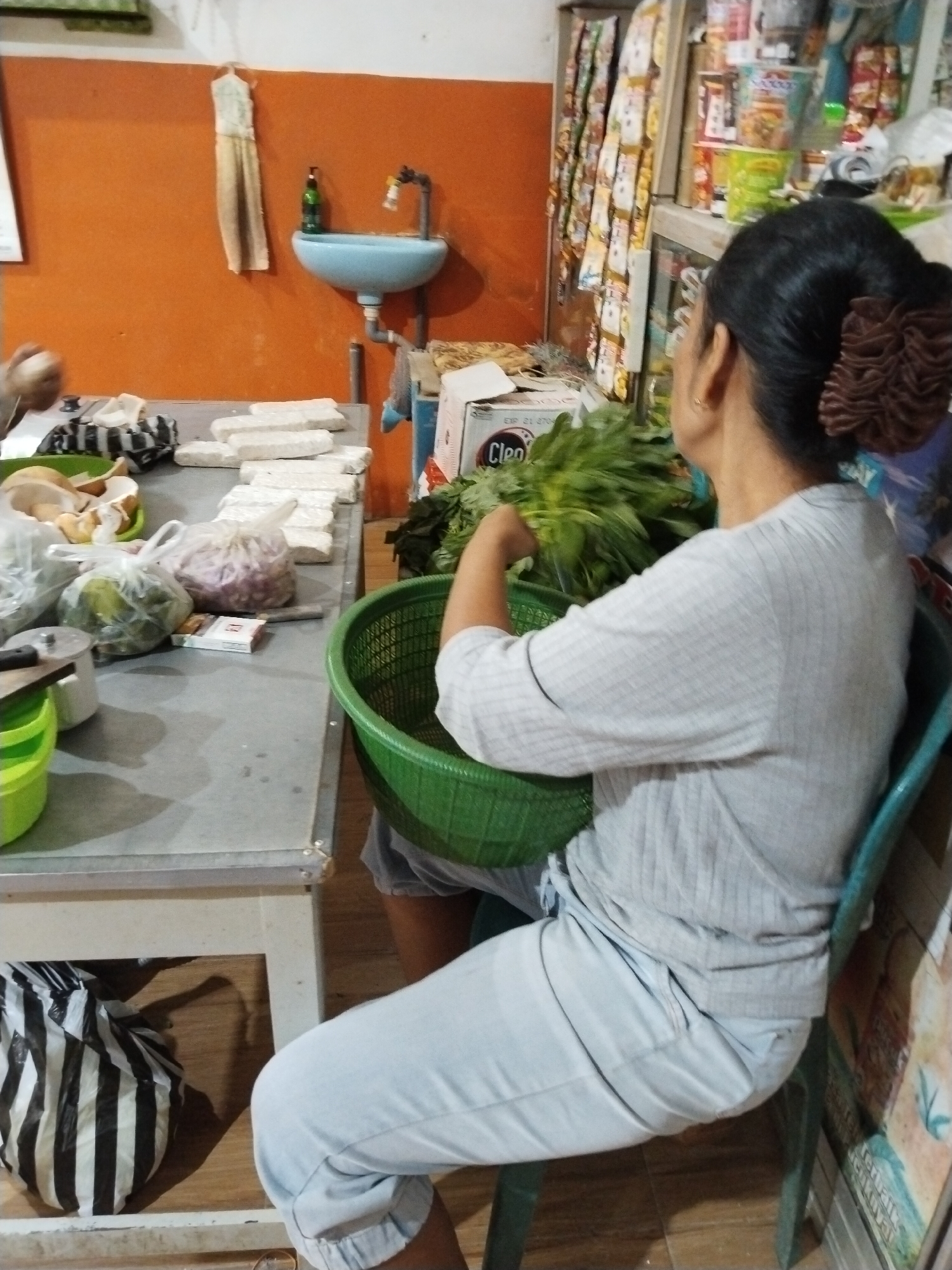A woman with her back to the camera, wearing a light gray long-sleeved shirt and light blue pants, sits on a turquoise chair in what appears to be a small shop or kitchen. Her dark hair is pulled up in a bun with a brown scrunchie. She is holding a large bunch of green leafy vegetables, possibly spinach or kale, which are placed inside a green plastic basket. To her left, a table is covered with various items, including several rectangular blocks of white food, likely tempeh, and plastic bags containing produce such as onions and green vegetables. A sink with a light blue basin is mounted on an orange wall in the background, with a bottle of green liquid and a faucet visible. Shelves in the background are stocked with packaged food items. A black and white striped bag is on the floor beneath the table. The overall setting suggests a food preparation or market environment in Prigi, Indonesia.