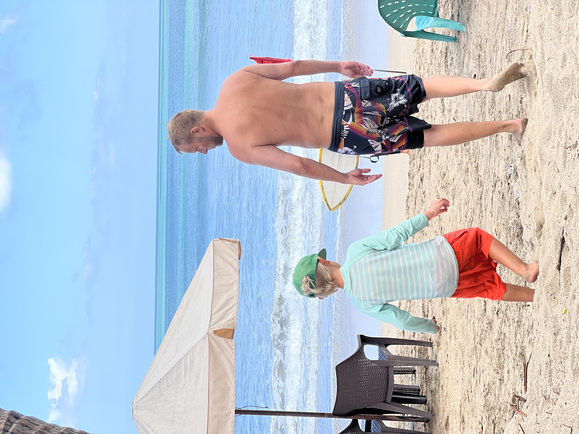 The image captures a beach scene in Kuta, Indonesia, on a sunny day with a clear blue sky. The perspective is from a low angle, looking upwards, and the image is rotated 90 degrees counter-clockwise.
In the foreground, a young child wearing a green cap and a light blue and white striped long-sleeved shirt with orange shorts is standing on the sand. The child appears to be looking down towards the sand or the water, with their right arm slightly extended.
Behind the child, a man with blonde hair, seen from the back and side, is walking on the beach. He is wearing colorful swim trunks with a black base and a tropical pattern. His arms are down by his sides, and he appears to be walking towards the water. A white surfboard with yellow trim is lying on the sand near him. A red flag is visible in the background.
The background shows the ocean with gentle waves and the distinct turquoise blue of the water characteristic of the area. On the sand, there are a few beach chairs, one of which is green and appears to be made of plastic, and another darker chair is partially visible. A portion of a large, light-colored beach umbrella is also visible in the lower left of the frame, suggesting a relaxed beach day. The sand itself is light-colored and appears to have some debris scattered on it, typical of a well-used beach. There is no visible text in the image.