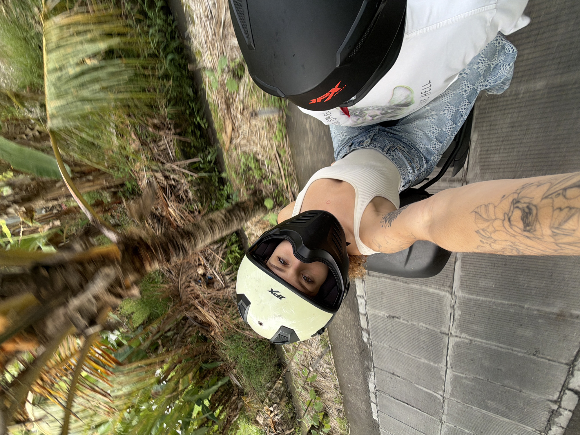 In North Kuta, Indonesia, a woman with curly blonde hair is riding on a motorcycle, wearing a cream-colored helmet with black accents and a white tank top. She is looking back over her shoulder at the camera, her eyes clearly visible. Her arm, adorned with tattoos, is extended towards the camera. She is wearing blue denim jeans with a white patterned overlay. The second person, whose arm is extended to take the photo, also has tattoos. They are wearing a black helmet with red accents. The motorcycle has a black seat and is moving along a paved road with textured concrete sections. The surrounding environment is lush and tropical, with palm trees and dense green foliage visible.