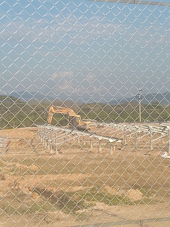 Behind a chain-link fence, a construction site is visible. An excavator, labeled "HYUNDAI," sits atop a metal framework with evenly spaced concrete supports. The ground is a mix of dry dirt and sparse vegetation. In the background, a green, hilly landscape meets a clear sky. A lone utility pole stands near the horizon. The overall scene conveys a sense of industrial development in a rural setting.