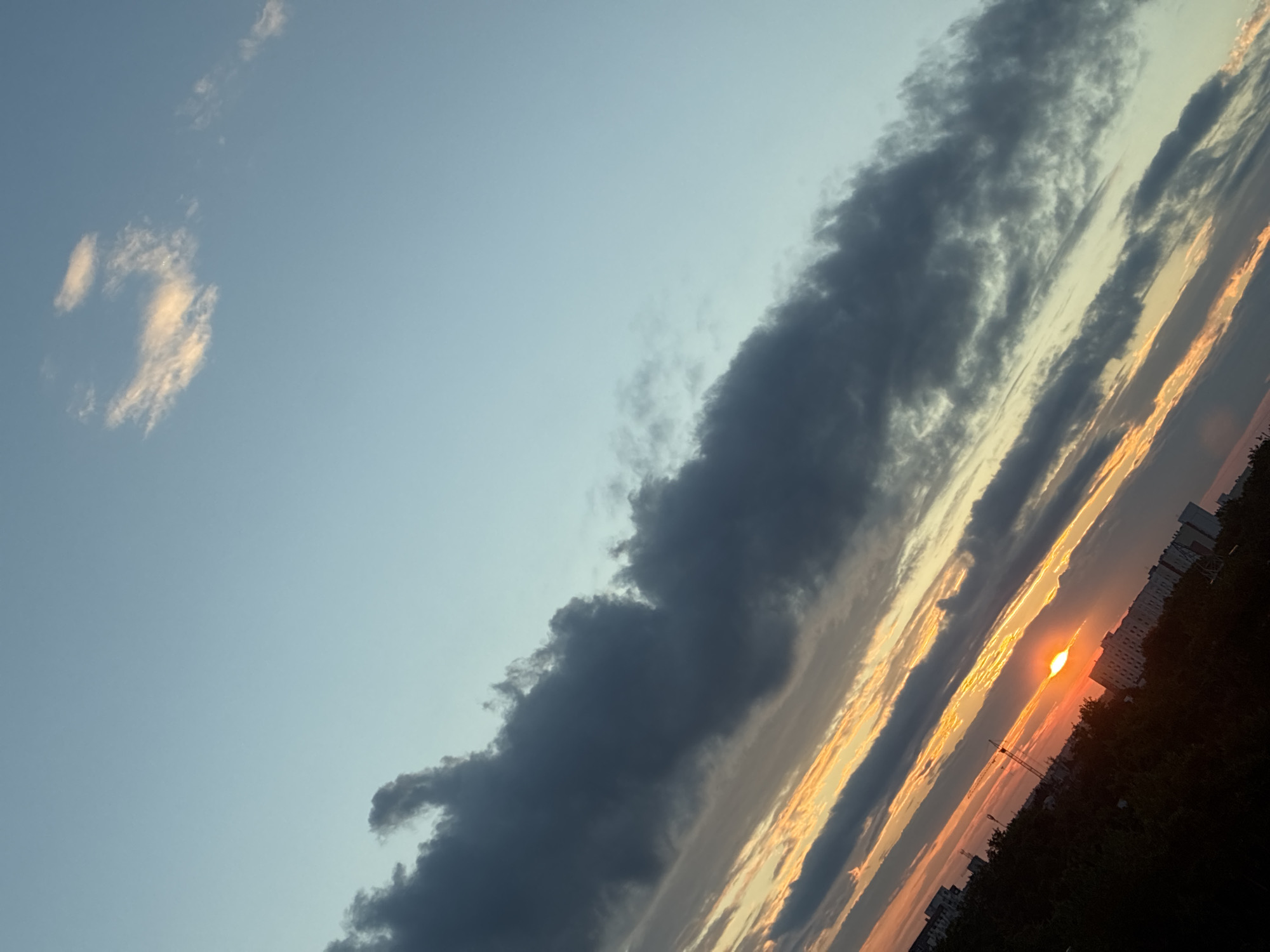 The image shows a tilted, low-angle view of a sky at sunset over a city. The sky is a gradient of pale blue at the top, transitioning to lighter blues and yellows closer to the horizon, with streaks of orange and pink from the setting sun. Dark gray clouds are scattered across the sky, some appearing wispy and others more dense. The sun itself is a bright orange orb partially obscured by clouds, located near the center of the lower half of the frame. In the foreground and bottom of the image, the dark silhouettes of trees and buildings are visible, including apartment buildings and what appears to be a construction crane. The scene is overall dark due to the sunset.