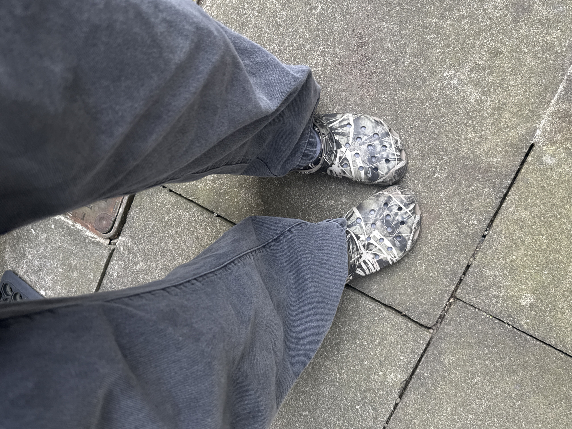 The image shows a first-person, downward-angled view of a person's legs and feet standing on a paved surface. The person is wearing loose-fitting, gray jeans and a pair of gray and white camouflage-patterned Crocs. The paved surface appears to be composed of gray stone tiles of varying shapes and sizes, with visible joints or seams between them. The setting seems to be a street or pedestrian area. The exact location within London, United Kingdom, cannot be determined from the visual evidence.