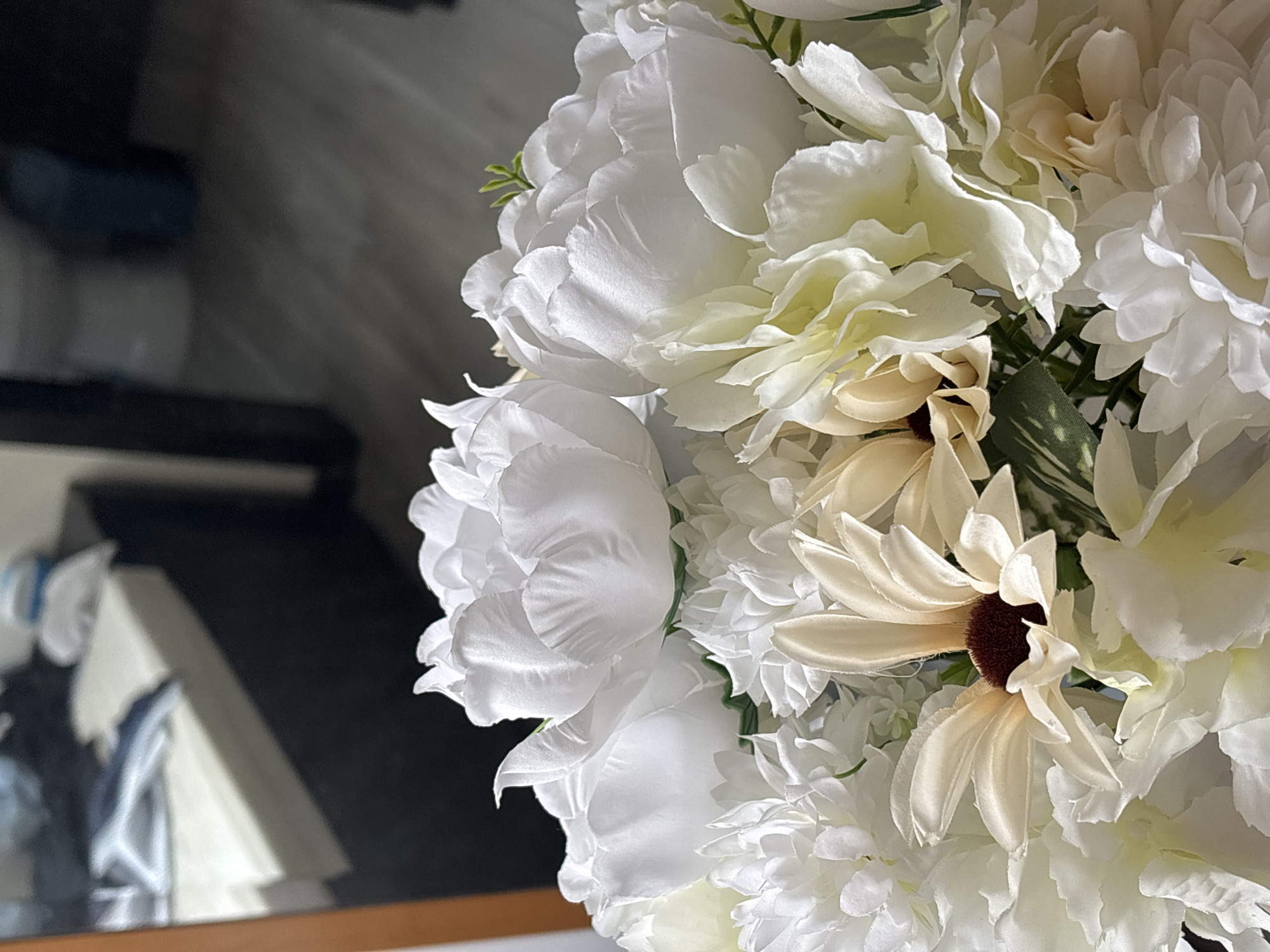 A close-up shot shows an arrangement of artificial white and beige flowers. The focus is on the floral details, with several types of flowers intermixed, including what appear to be peonies and daisies. Some of the flowers have dark brown centers. A blurry reflection of a television and a white object is visible in the dark background on the left side of the frame. No specific information can be derived about the location of Santa Clara Ocoyucan, Mexico, or the people, subjects, objects, actions, or interactions within the environment, other than the flowers in the foreground.
