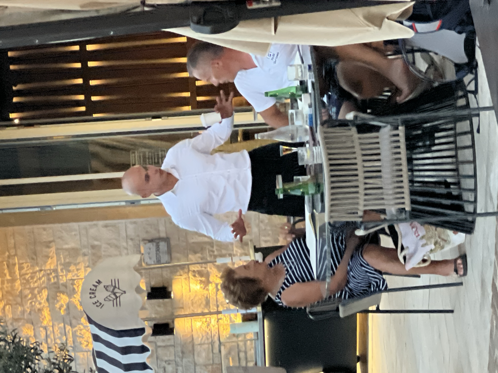 The image depicts an outdoor seating area of a restaurant or cafe in Đenovići, Montenegro. A male server, wearing a white collared shirt and black pants, is interacting with two patrons seated at a round table. The server is holding a white cup and gesturing with his right hand.
To the left of the server, a woman with short, curly brown hair is seated at the table, wearing a black and white striped dress. She is looking towards the server.
To the right of the server, a man is seated at the table, wearing a white polo shirt. He is also looking towards the server and appears to be speaking.
The table is set with glasses, bottles of water and what appears to be a bottle of alcohol. There are also cigarettes on the table.
In the background, there is a building with stone walls and vertical lighting fixtures. A striped awning with the words "ICE CREAM" is visible on the left side of the frame. The overall lighting suggests it is late afternoon or early evening.
