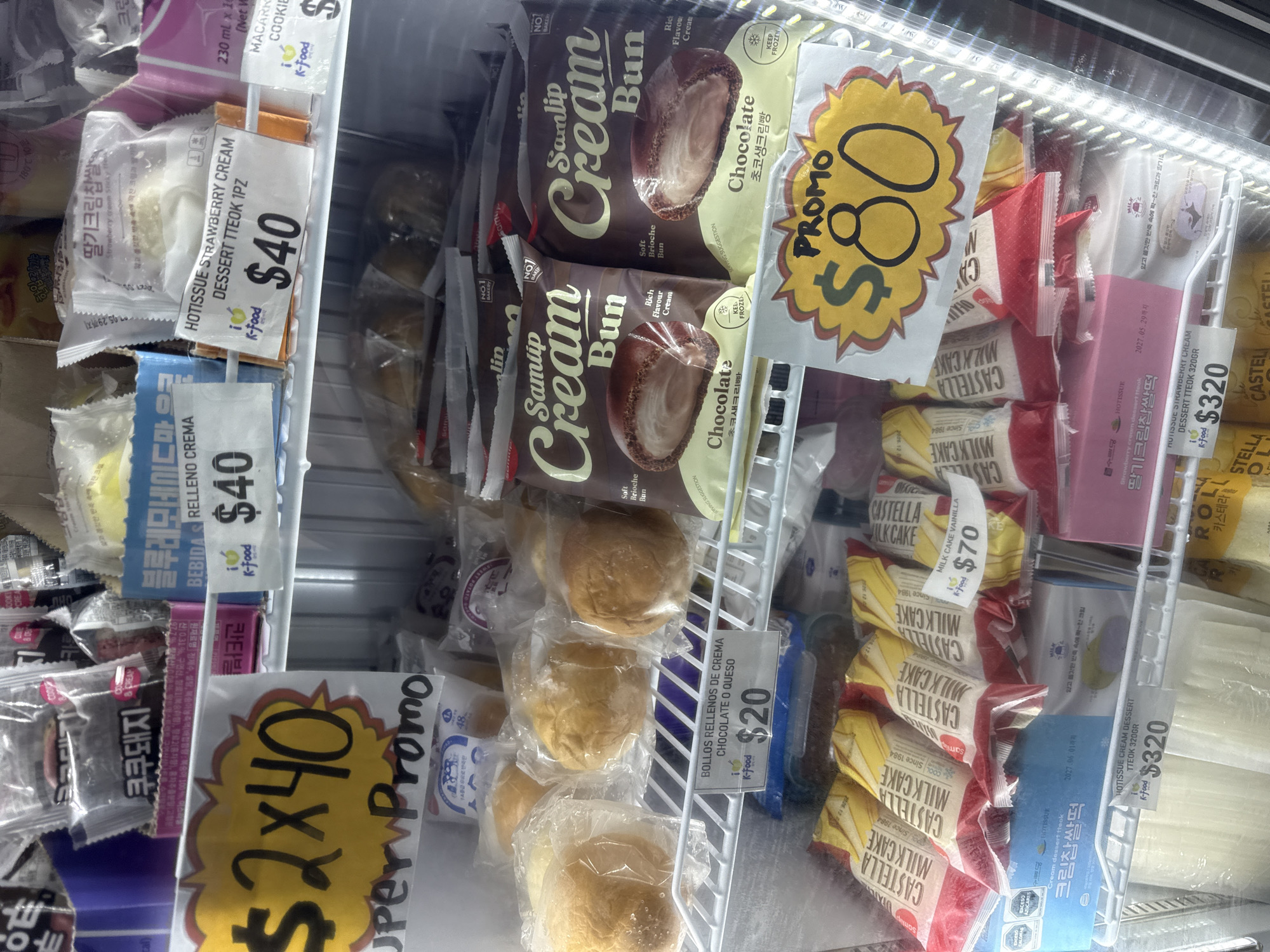 This image displays the interior of a brightly lit refrigerated display case in a store, likely an Asian grocery store given the product selection and languages. The shelves are stocked with various packaged food items, predominantly Korean and some with Spanish labels, indicating an international market setting in San Andrés Cholula, Mexico.
The top shelves feature multiple packages of "Samlip Cream Bun Chocolate" (초코생크림빵), described as a "Soft Brioche Bun" with "Rich Flavour Cream," priced with a "PROMO $80" sign. Below these, there are several individual cream-filled buns (likely "BOLLOS RELLENOS DE CREMA CHOCOLATE O QUESO") in clear plastic, priced at "$20" each.
On the left side, there are "HOTISSUE STRAWBERRY CREAM DESSERT TTEOK" (딸기크림찹쌀떡) priced at "$40" for a single piece, and a "BEBIDAS RELLENO CREMA" (cream-filled drink) also for "$40". A "Super Promo $2x40" sign is visible near a product with the text "쿠쿠돼지". Larger boxes of "HOTISSUE CREAM DESSERT TTEOK 320GR" (크림찹쌀떡) are priced at "$320".
The right side of the case holds stacks of "CASTELLA MILK CAKE" (카스테라), with a "MILK CAKE VAINILLA" variant priced at "$70". Many products feature the "K-Food" logo, and some have visible expiration dates like "2027.05.29까지" and "2027.06.01까지". The overall scene suggests a well-stocked section for frozen or chilled desserts and snacks.