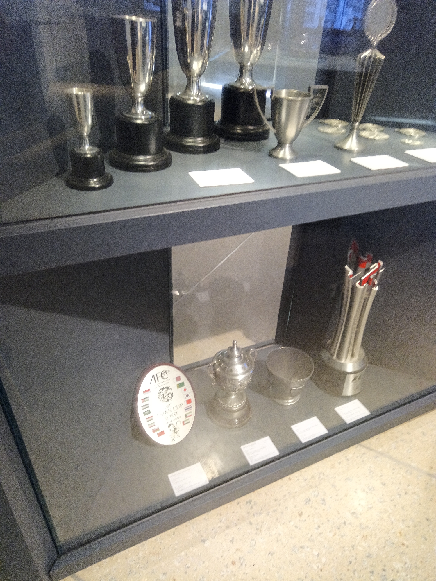 A glass display case contains a collection of silver trophies. From left to right, there are three tall, slender trophies with black bases, followed by a small goblet-style trophy. To the right of these are a more ornate, smaller trophy with handles, and a bucket-shaped trophy. Further to the right, a large, multi-pronged trophy with red accents stands prominently. In the foreground, to the left, a circular plaque with the "AFC ASIAN CUP" logo and various flags is visible. The display case is set against a dark gray background. The floor beneath the display case appears to be a light-colored terrazzo. This scene is likely located at the Asian Football Confederation (AFC) headquarters in Kuala Lumpur, Malaysia, where historical AFC trophies are displayed.