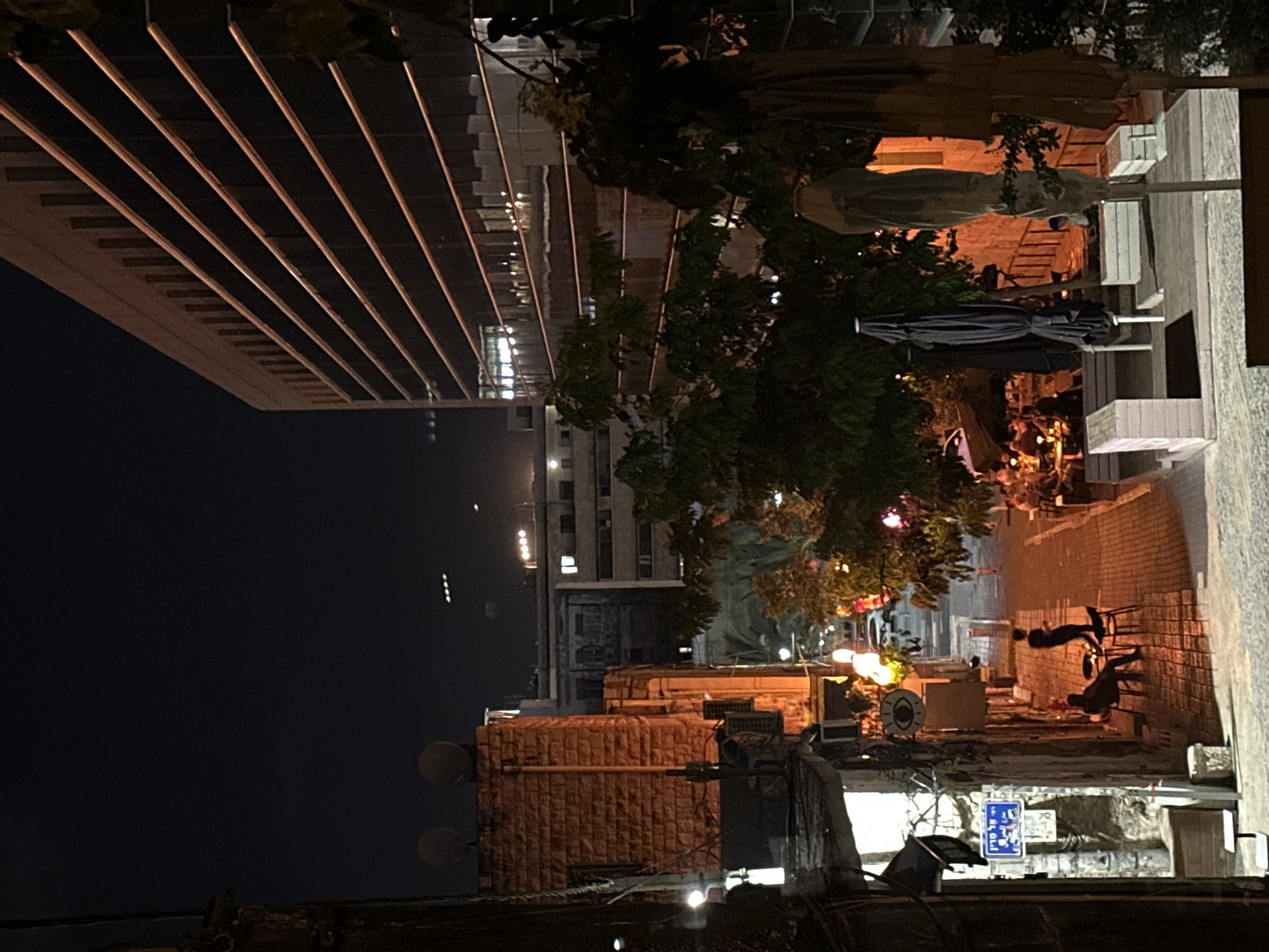 This image depicts an outdoor scene at night, likely in Haifa, Israel. The perspective is tilted, giving a sense of looking from a slightly elevated position down a street or alleyway.
In the foreground, there are remnants of urban infrastructure, including what appears to be a wall or ledge with some piping and lights. Further back, a brick building is visible, adorned with decorative lighting and what look like satellite dishes. Above this, a modern building with a glass facade and parallel horizontal lines can be seen.
The midground features a tree with festive, colorful lights, suggesting a gathering or celebration. Nearby, there are several dark umbrellas and what might be outdoor seating, hinting at a restaurant or café. Further down the alley, two figures are visible, silhouetted against the light, seemingly engaged in conversation or moving.
The overall atmosphere is nocturnal and somewhat lively due to the lighting. The presence of what appears to be a street sign with Hebrew text indicates an urban setting in Israel. The sky is dark, confirming the time of day. The image captures a slice of urban life at night, with a mix of modern and older architecture, and hints of social activity.