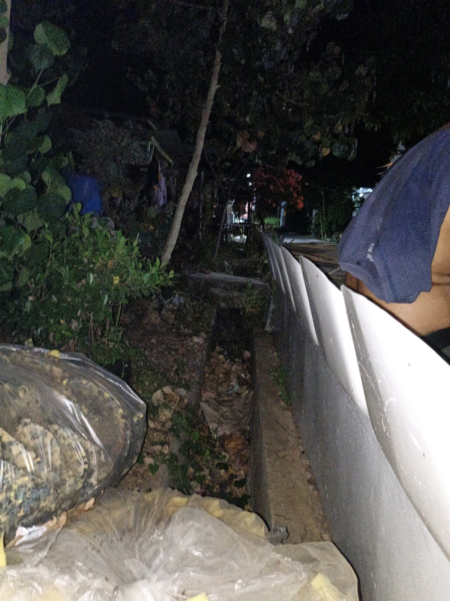 The image shows a cluttered outdoor scene at night, likely in Cikal, Indonesia. In the foreground, there are large plastic bags filled with what appears to be shredded material or debris. To the right, a person is partially visible, wearing a dark blue t-shirt, and leaning over a white, textured wall or barrier. The background features dense vegetation, trees, and hints of structures, suggesting a semi-urban or residential area. A narrow concrete channel or ditch runs along the base of the white barrier, filled with debris and dirt. The overall impression is one of an unkempt, outdoor space at night, possibly near a roadside or alleyway, with the presence of discarded materials and a person.