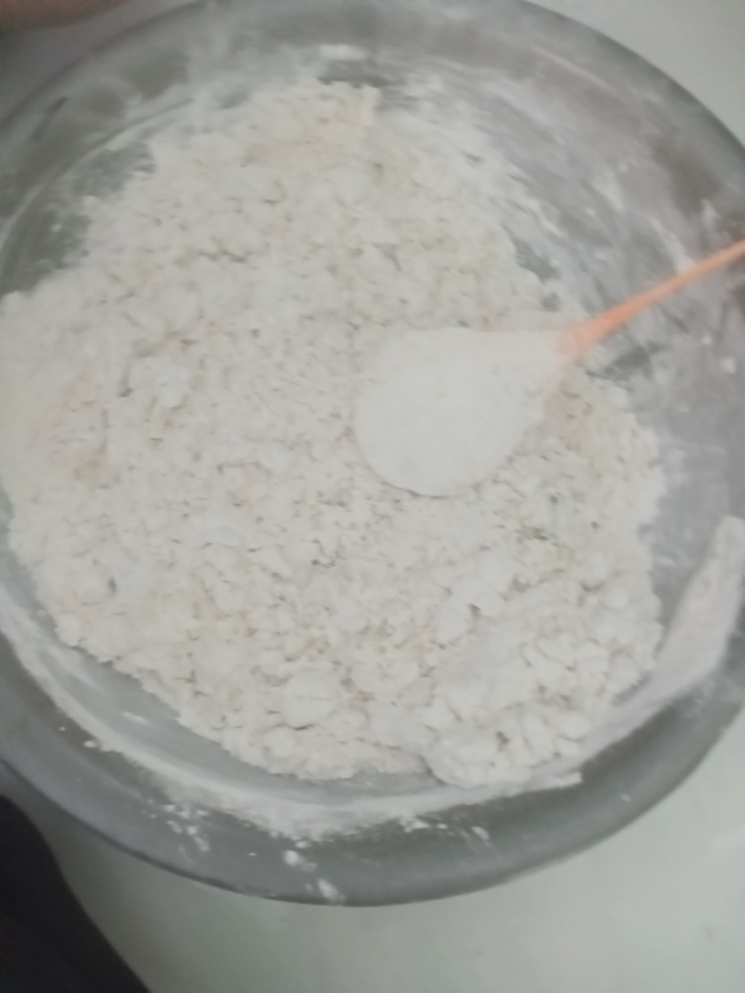 In a clear glass bowl, white flour is visible, with a small orange spoon partially submerged in the flour. The scene appears to be indoors, likely in a kitchen or food preparation area, but there is no specific information available to pinpoint its location within Badas, Indonesia. The lighting is soft and diffused, casting gentle shadows. The overall impression is one of a simple, everyday activity related to cooking or baking.