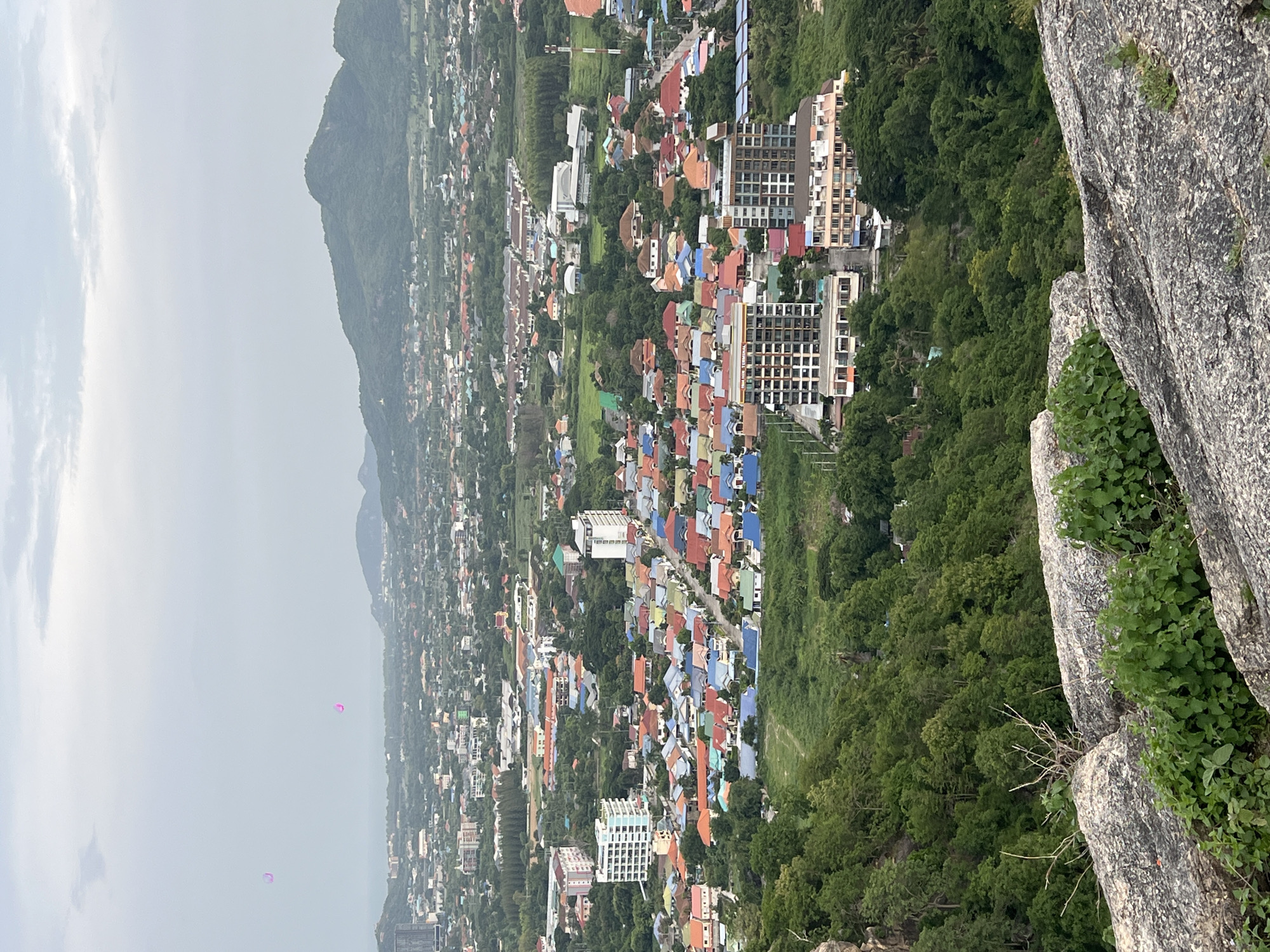 An elevated, wide-angle shot captures the cityscape of Hua Hin City Municipality, Thailand, under a hazy, overcast sky. In the foreground, large, textured grey rocks covered in patches of green vegetation dominate the bottom of the frame. Beyond the rocky outcrop, a lush green hillside slopes down towards the densely packed urban area. The city is characterized by a mix of buildings: rows of colorful residential homes with red, blue, and orange tiled roofs are clustered together, interspersed with taller, modern apartment buildings and other commercial structures. A hazy horizon reveals distant, rolling green mountains and the faint line of the sea. Two small, indistinct pink or purple objects, possibly balloons or soap bubbles, are visible floating in the sky on the left side of the image. The overall impression is a sprawling, verdant city nestled against a natural landscape.