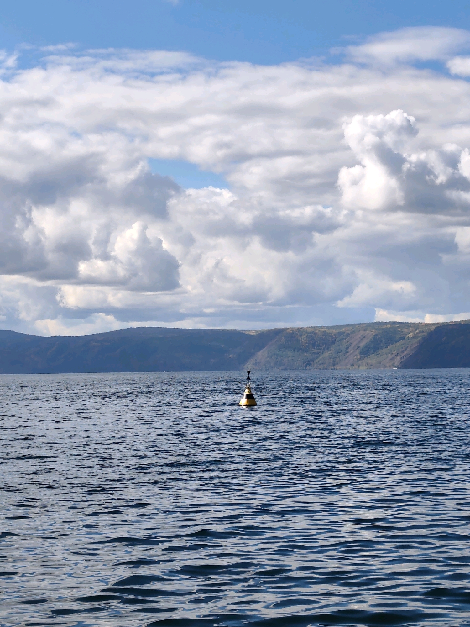 The image shows a wide view of a calm, deep blue body of water, likely Lake Baikal, with gentle waves rippling across the surface. In the middle distance, a solitary buoy, black and yellow, floats on the water. Beyond the water, a distant, tree-covered mountain range stretches across the horizon, its slopes a mix of dark green and brown. The sky above is a bright blue, dotted with fluffy white and gray clouds, some of which are large and dramatic. The scene suggests a peaceful, natural environment.