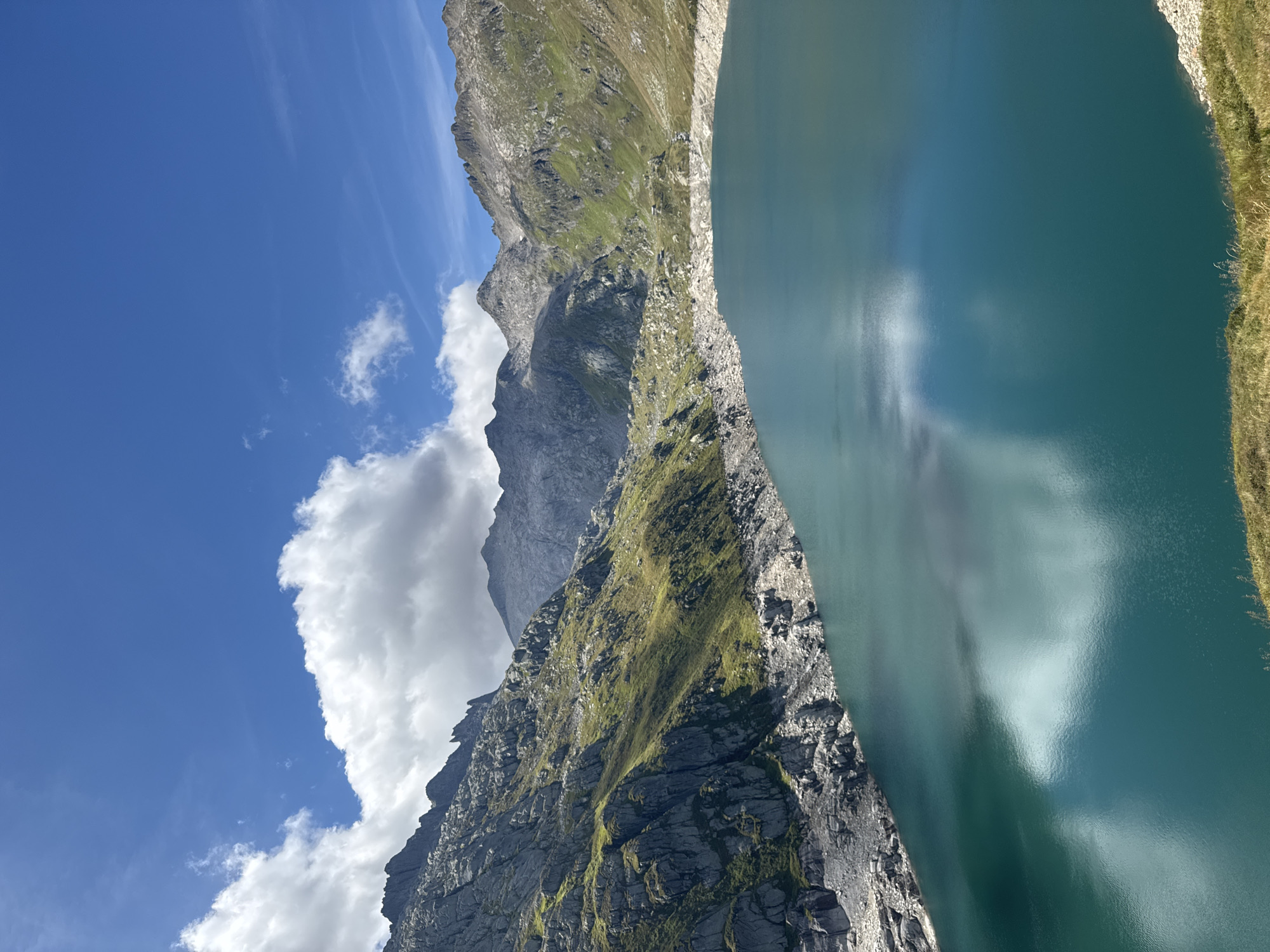 This is a photograph of a turquoise lake nestled amidst rocky, green mountains under a bright blue sky with scattered clouds. The lake's surface reflects the sky and the surrounding landscape. The shoreline is rocky, with sparse vegetation. The mountains rise steeply from the lake, with some areas covered in green grass and others appearing as bare rock. The scene is indicative of a high-altitude alpine environment. Based on the visual characteristics, this location is likely the artificial lake of the Ritom Dam, situated in the municipality of Quinto, near Airolo, Switzerland, within the canton of Ticino. There are no people or specific man-made structures immediately visible in the foreground or on the lake itself.