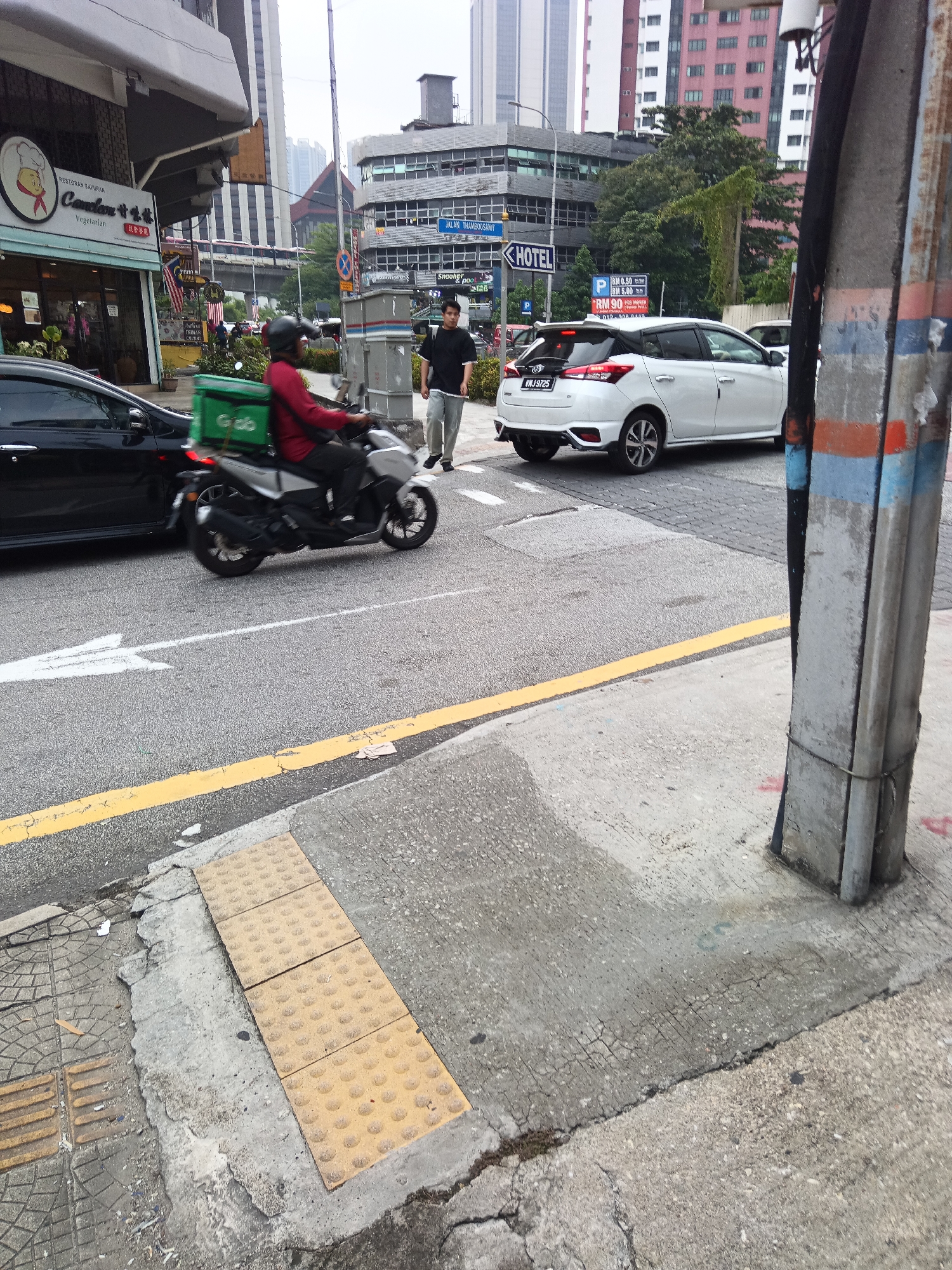 The image captures a street scene in Kuala Lumpur, Malaysia. In the foreground, a motorcycle with a green Grab delivery box is in motion, ridden by an individual in a red jacket and black helmet. To the left, a black car is also moving. In the background, a white hatchback car is stopped at an intersection, facing right. A man in a black t-shirt and grey pants is walking on the sidewalk to the right of the white car. Buildings of varying heights, including a modern high-rise and an older structure with visible signage, dominate the background. A traffic light and street signs are also present. The ground consists of asphalt roads and concrete sidewalks, with tactile paving visible near the curb. The overall environment suggests a busy urban setting.