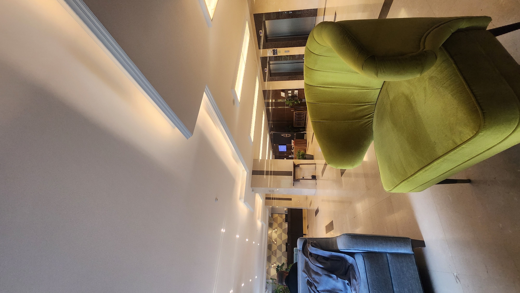The image depicts the interior of a hotel lobby. A bright green velvet sofa dominates the right side of the frame. In the background, elevators are visible. There is another sofa near the bottom edge of the image, gray-blue with a garment draped over it.
While the image does not contain explicit information about its location, it cannot be confirmed to be in Plovdiv, Bulgaria, without additional context.